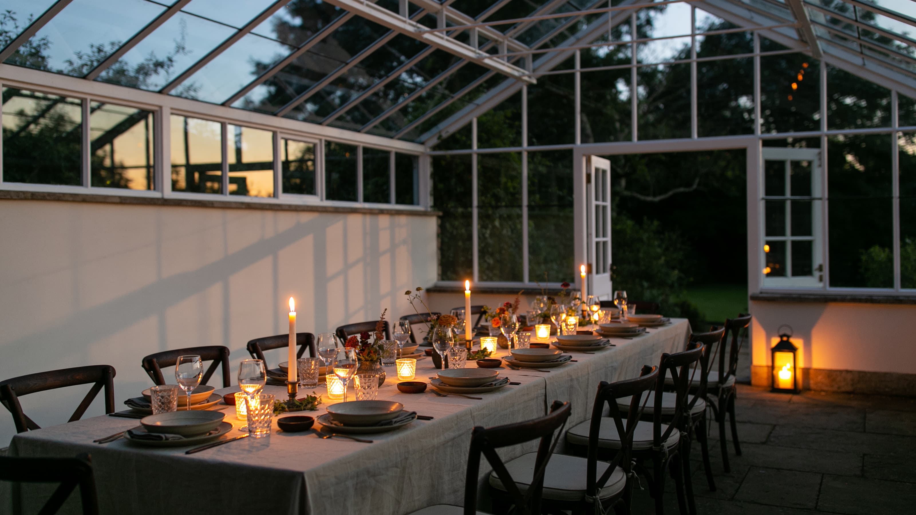 A long table set with candles inside a glasshouse at dusk, its doors open to a dark garden.