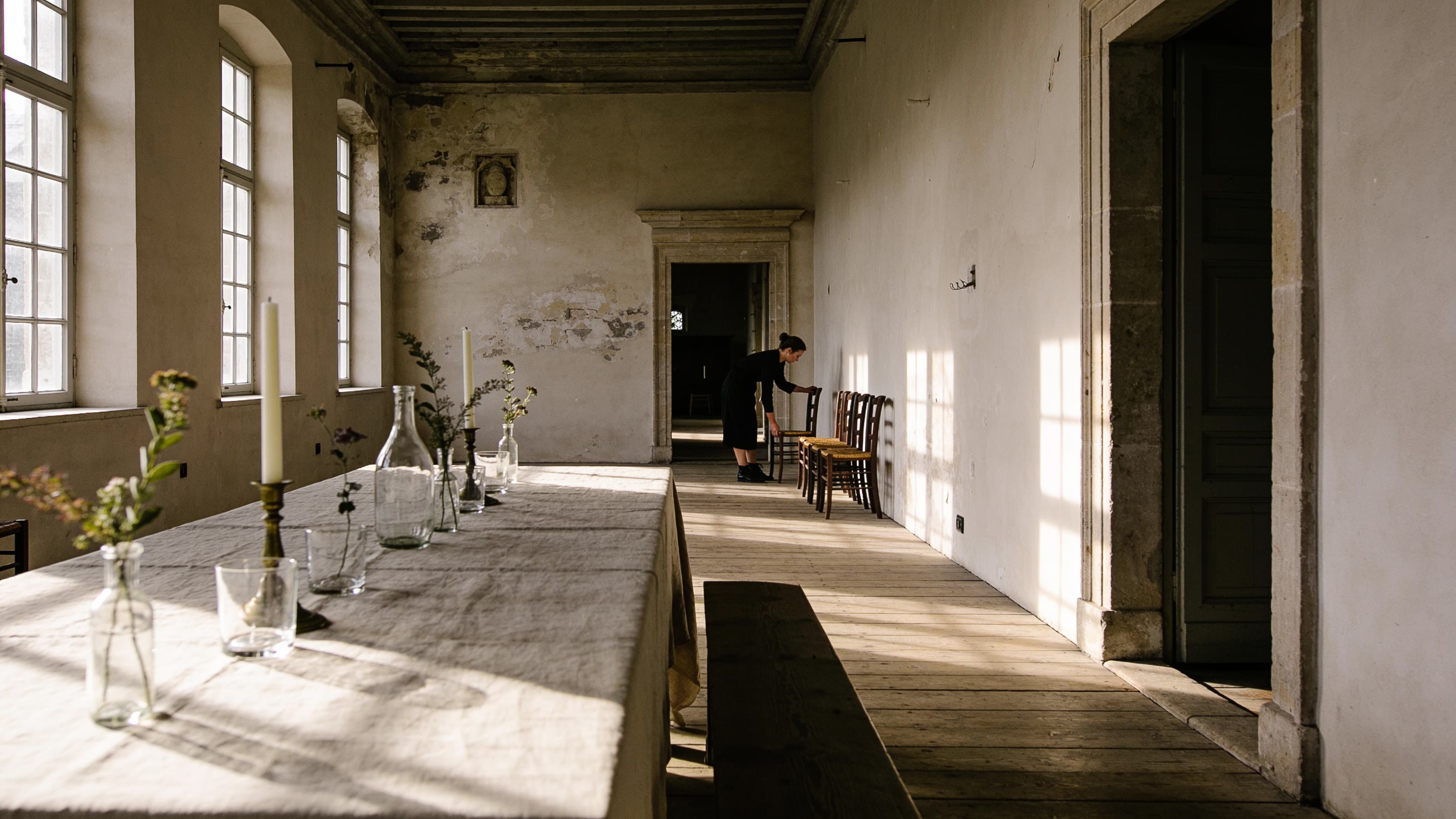 A long stone gallery in low afternoon sun: a linen-covered table beside tall windows, and a woman setting chairs against a pale plaster wall.