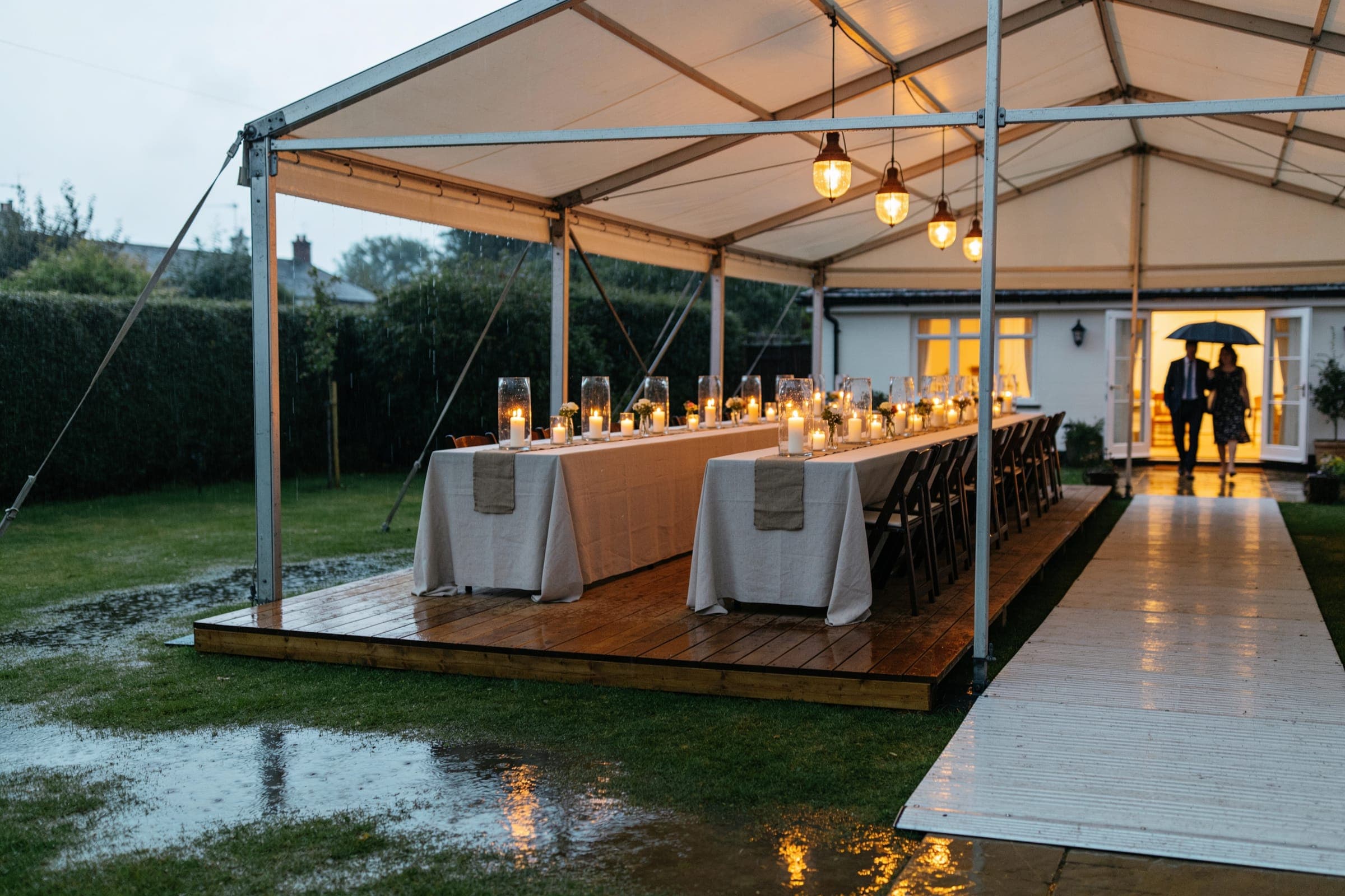 An open-sided marquee in a back garden in the rain, candlelit tables on a raised wooden floor and a boarded path to the house.