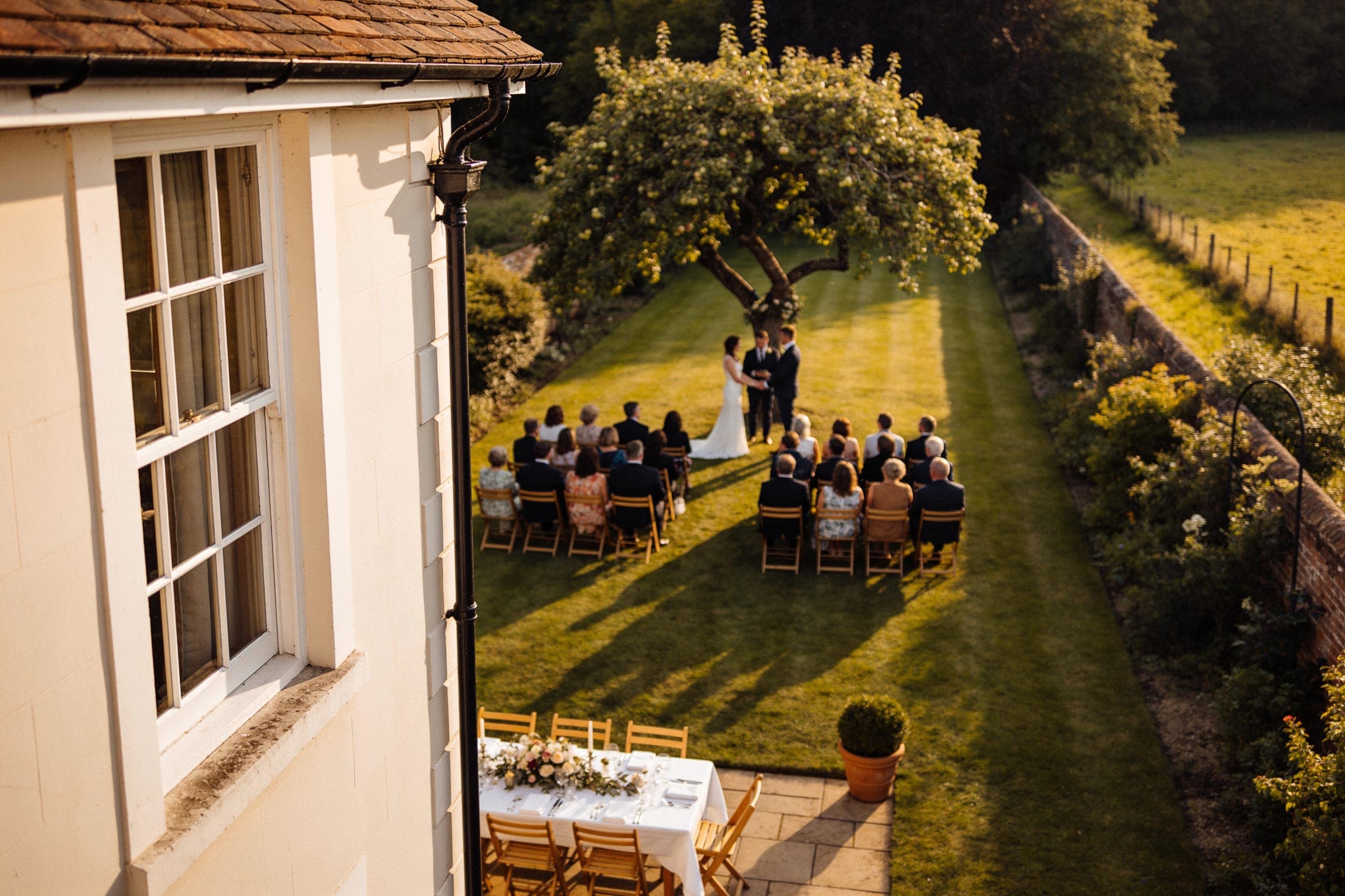 A small ceremony under an apple tree at the far end of a long garden, seen from the house, with a dinner table already set on the terrace below.