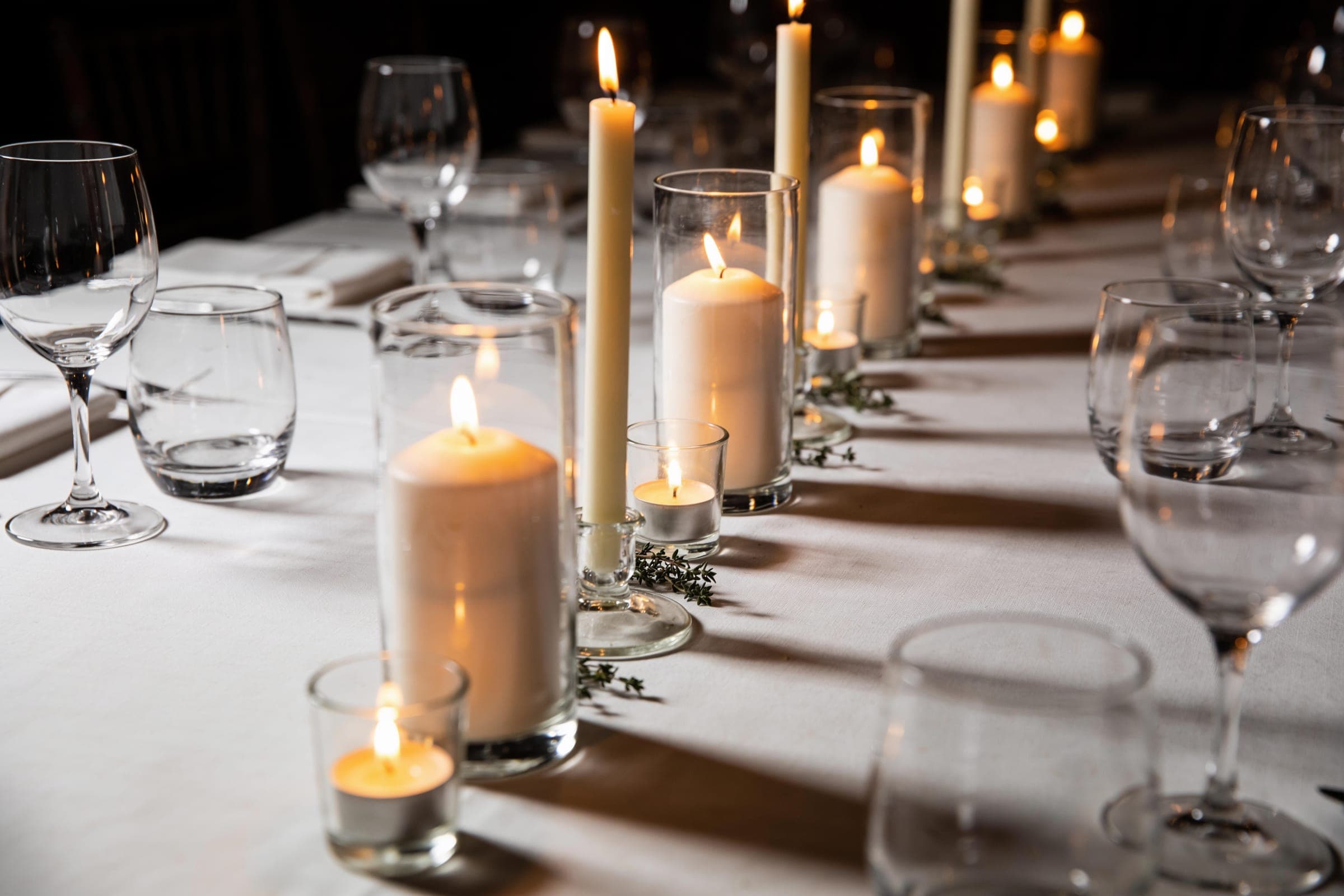 A line of pillar, taper and tea-light candles in clear glass of different heights down the center of a white table after dark.