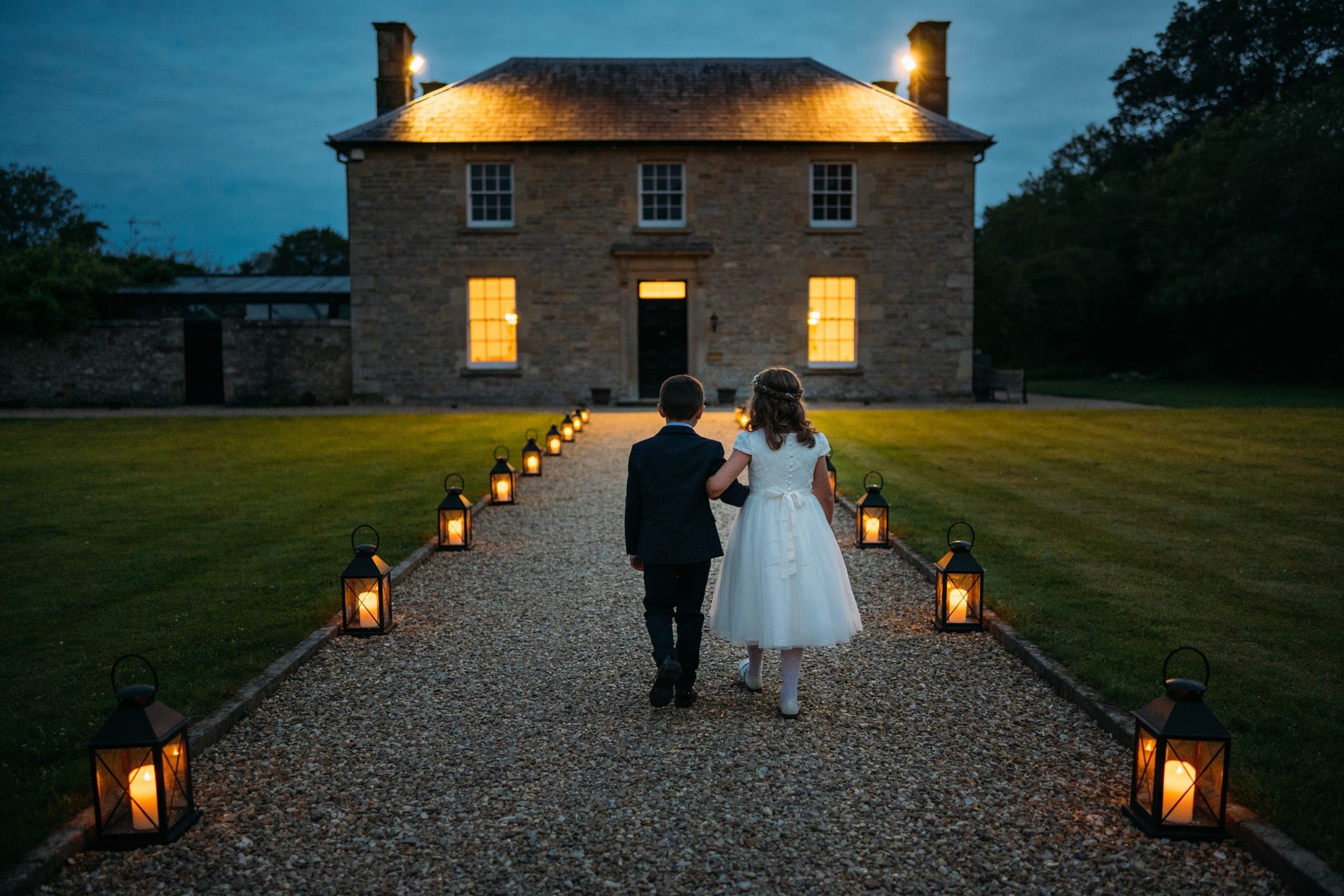 Two children walking arm in arm up a gravel path lined with lanterns toward the lit doorway of a stone house at dusk.