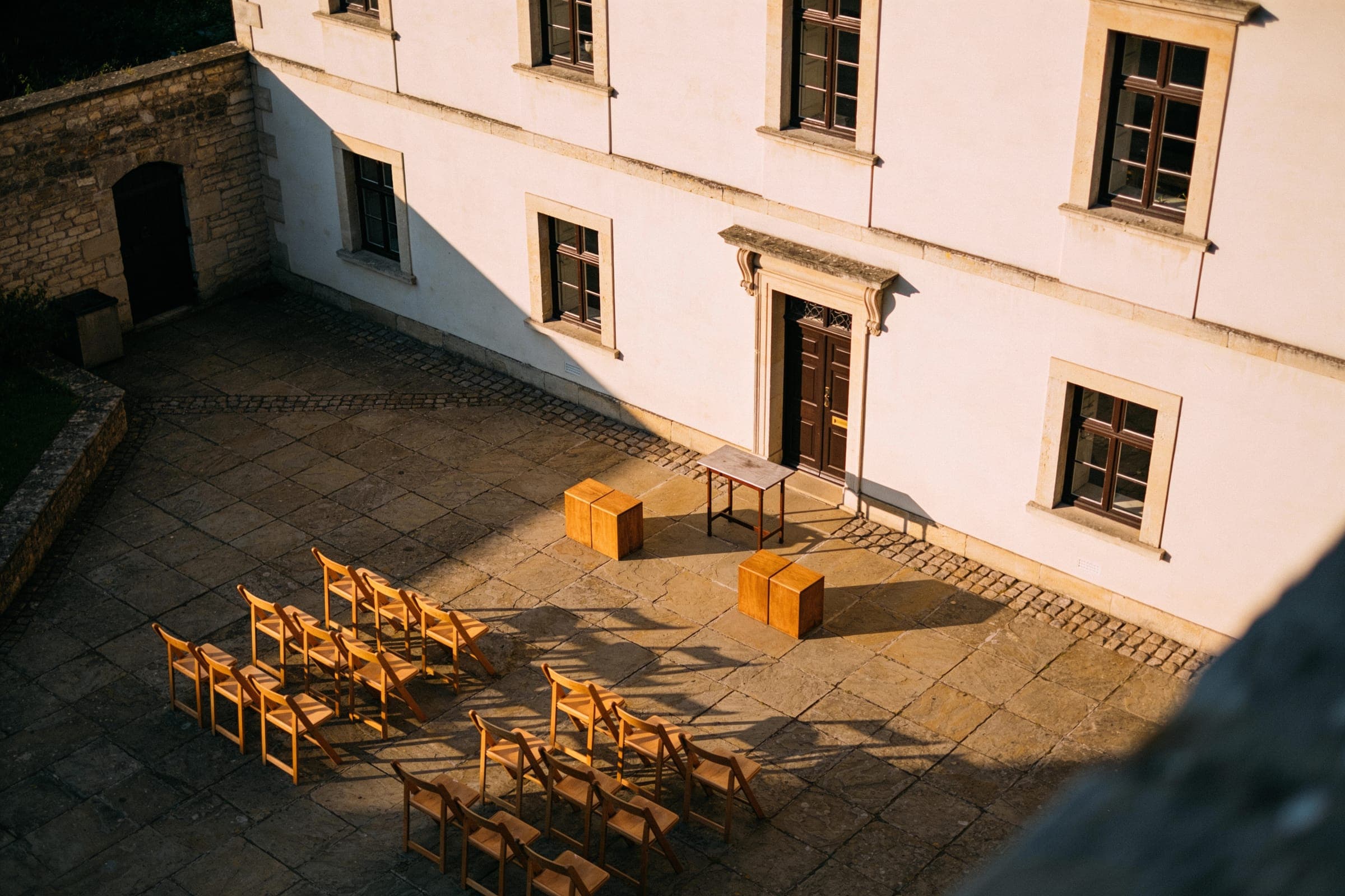 A paved courtyard seen from above in late-afternoon sun, with two short blocks of folding chairs set for a ceremony and the building's shadow on the wall.