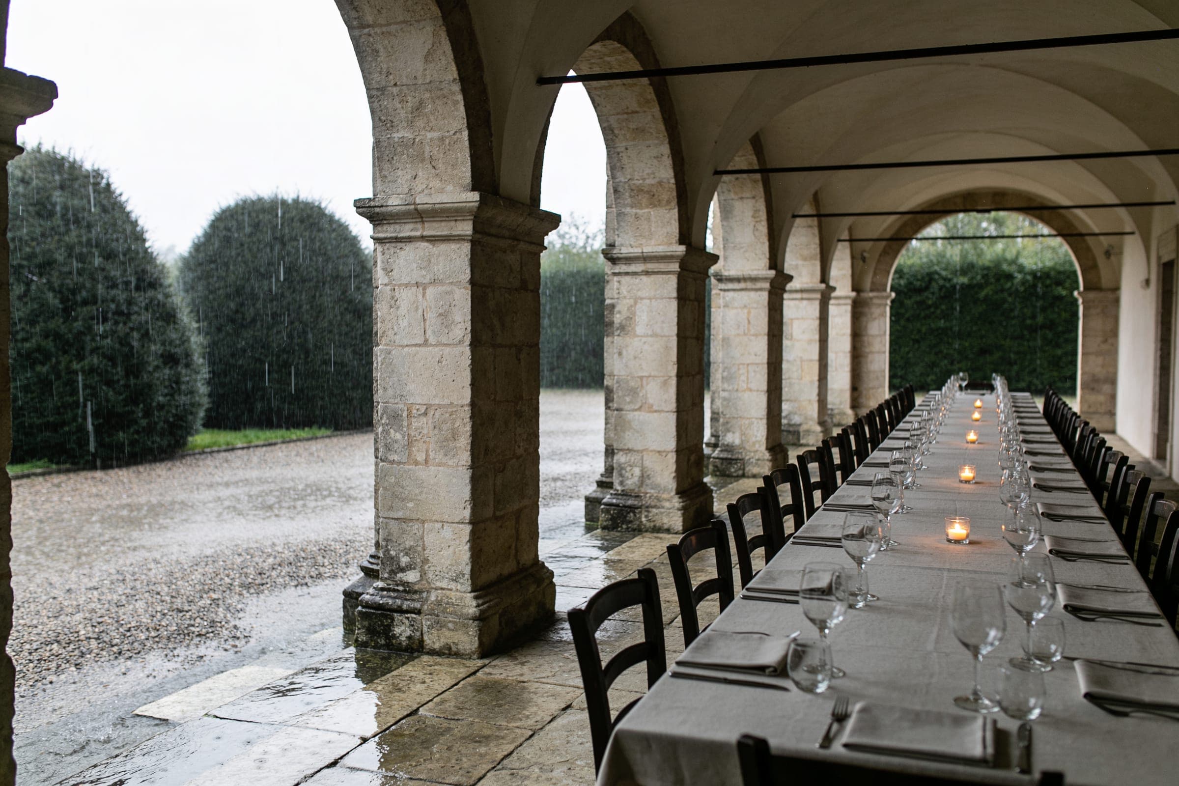 A long dinner table set under a stone arcade while rain falls on the gravel and clipped hedges beyond the arches.