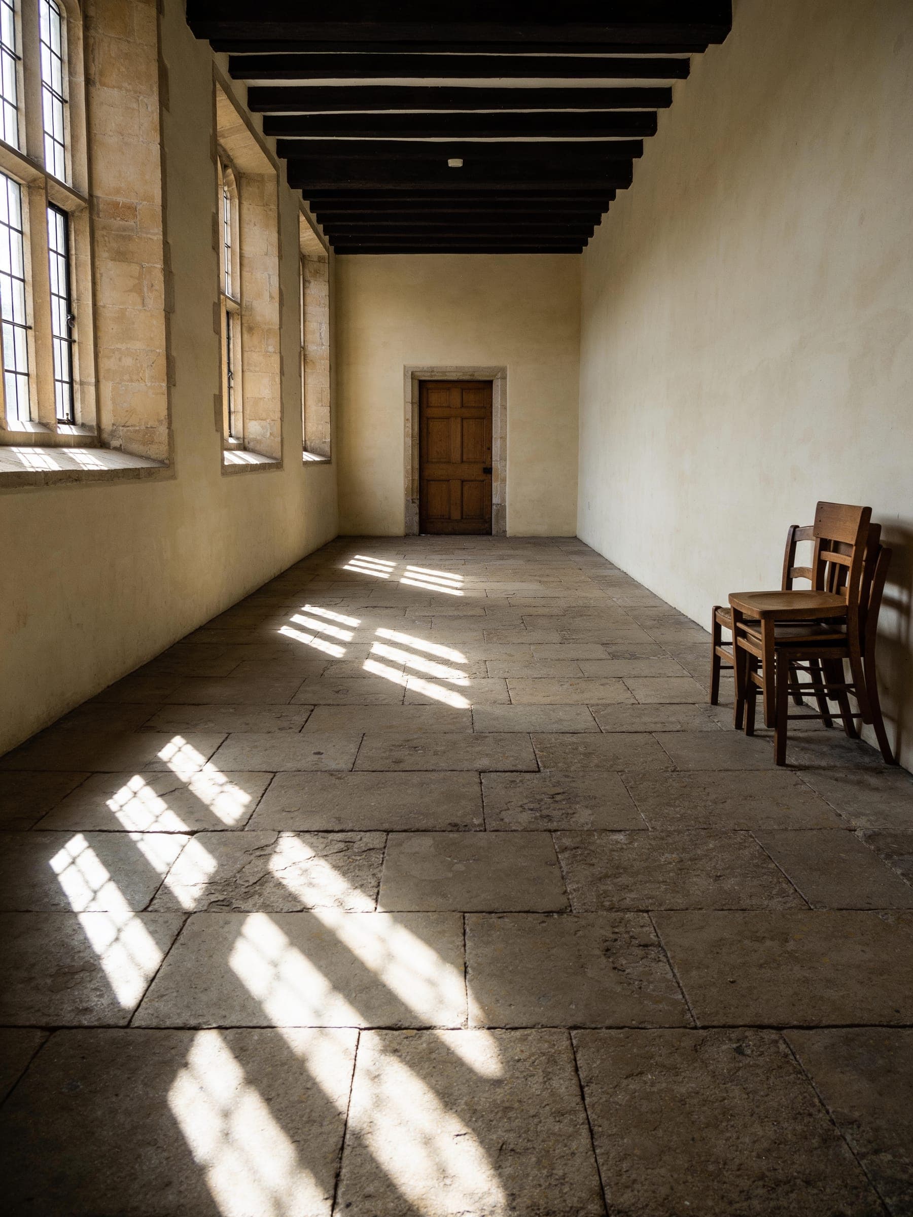 An empty stone-floored hall with tall windows along one side, sunlight falling across the floor and a few wooden chairs stacked by the wall.
