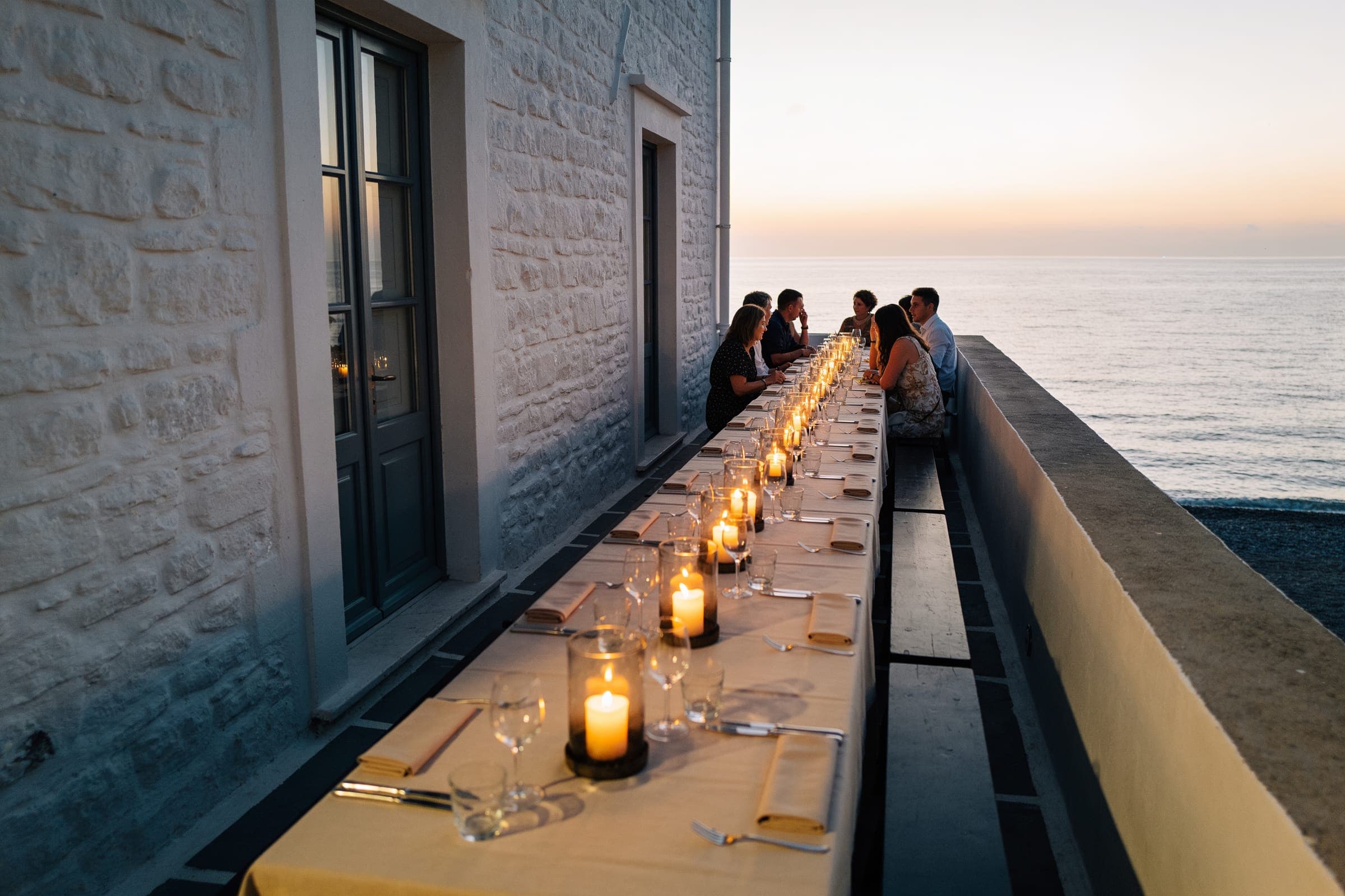 Guests at a long candlelit table set along the wall of a whitewashed building above the sea at dusk.