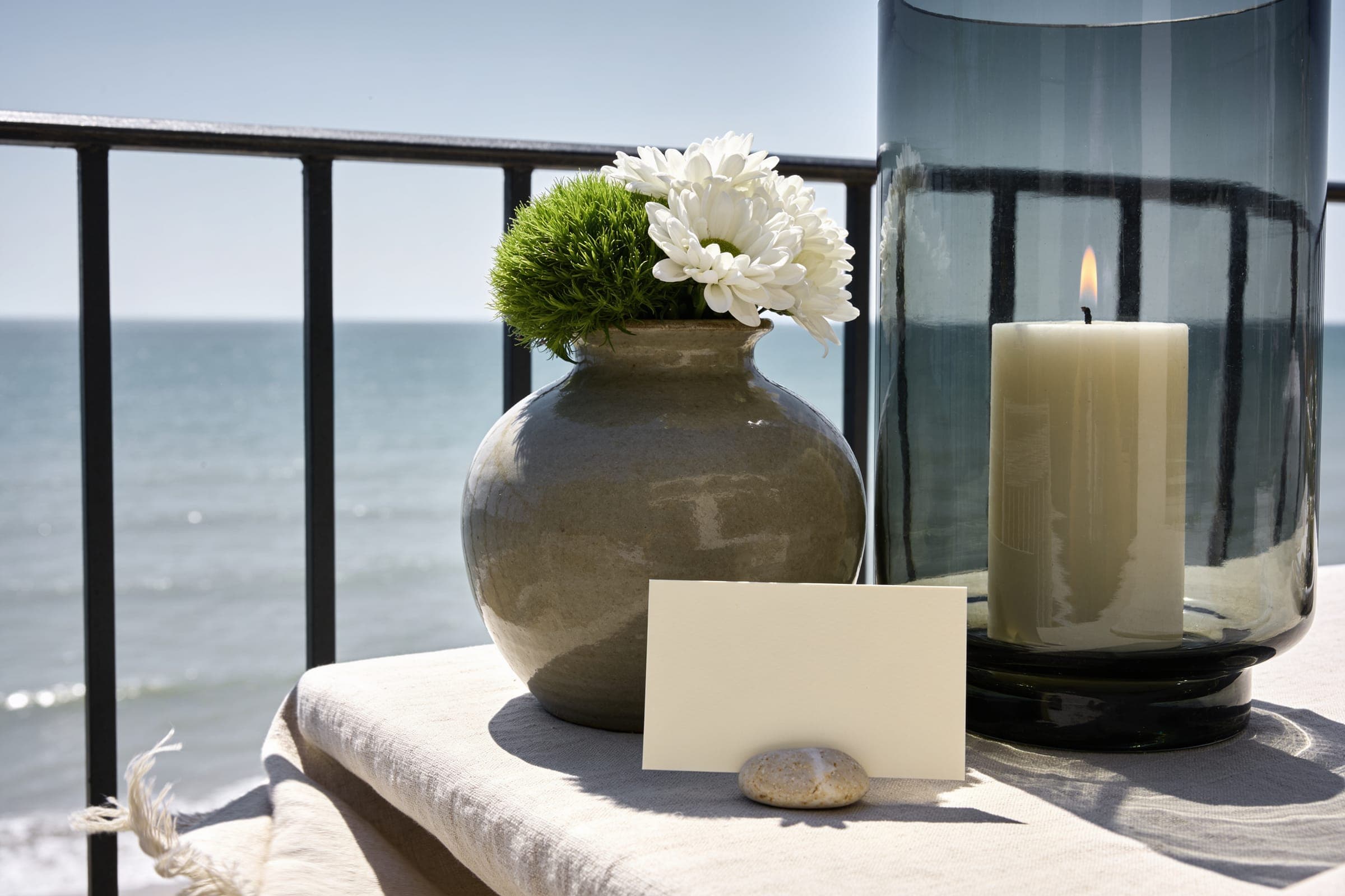 A heavy stoneware vase of white flowers, a candle inside a deep glass hurricane and a blank card resting against a small stone, on a terrace by the sea.