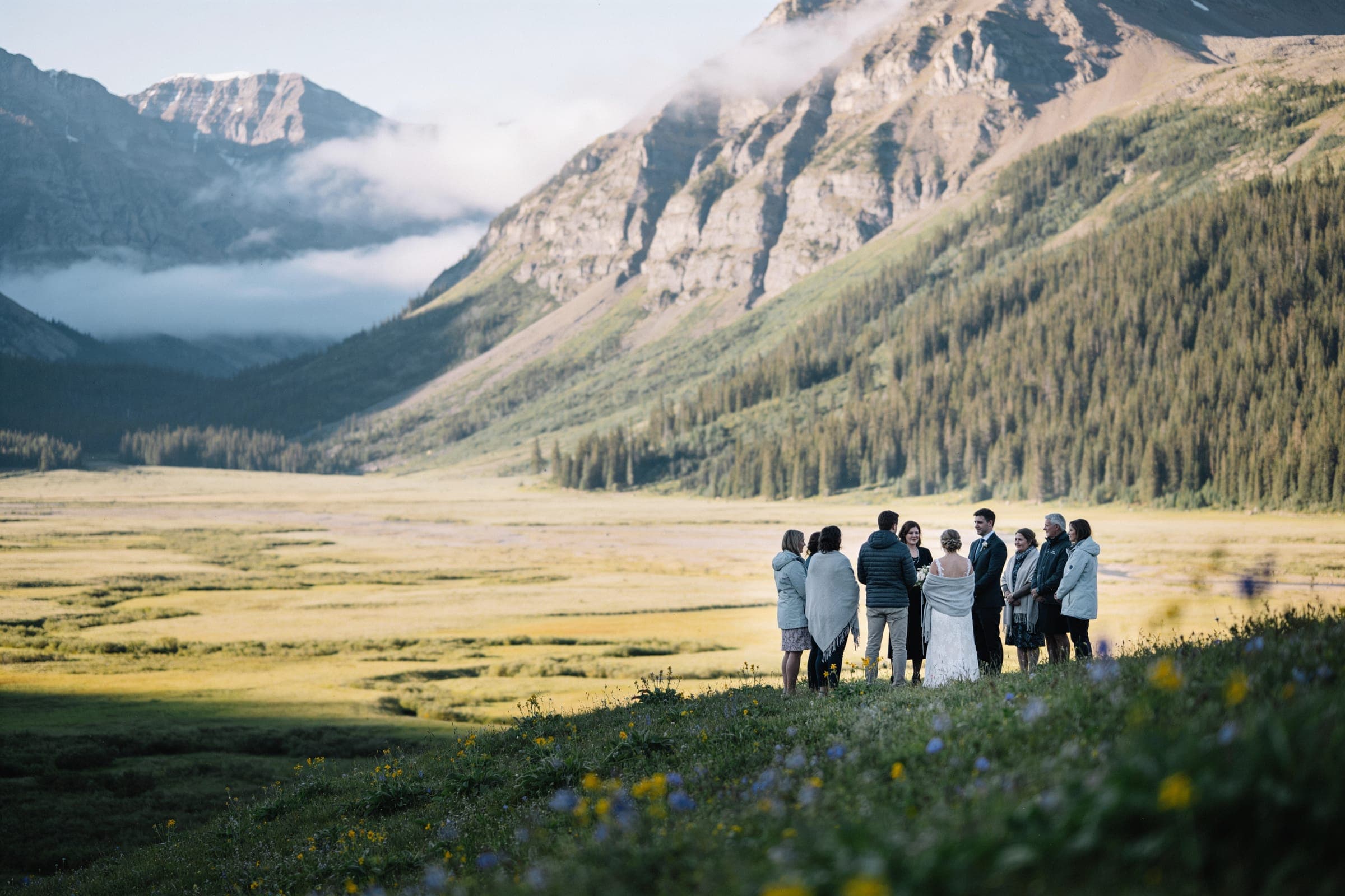 A small group standing around a couple on a wildflower slope above a wide mountain valley, cloud on the peaks.