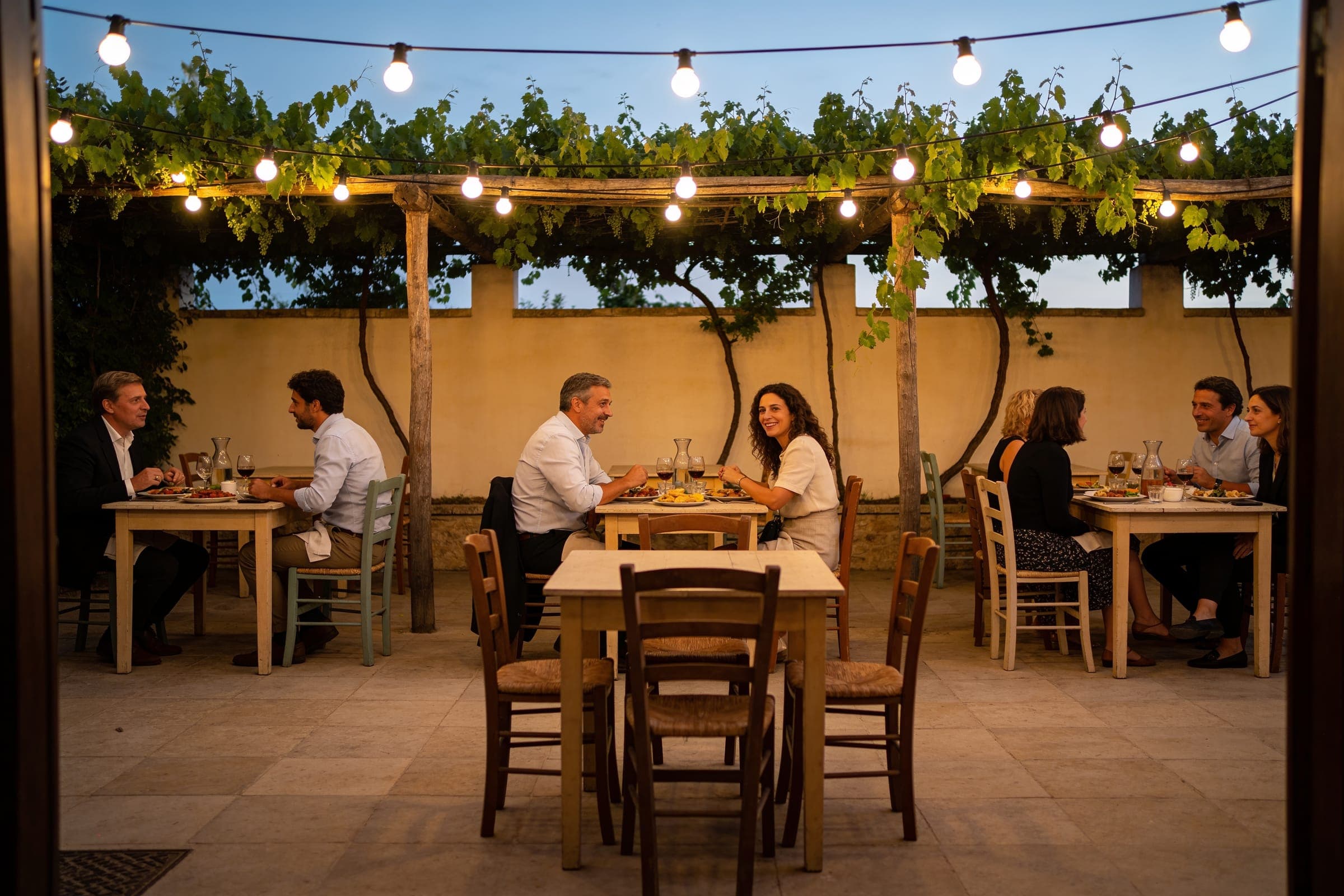 Guests at simple tables in a vine-covered courtyard under strings of bulbs, eating and talking in the evening.