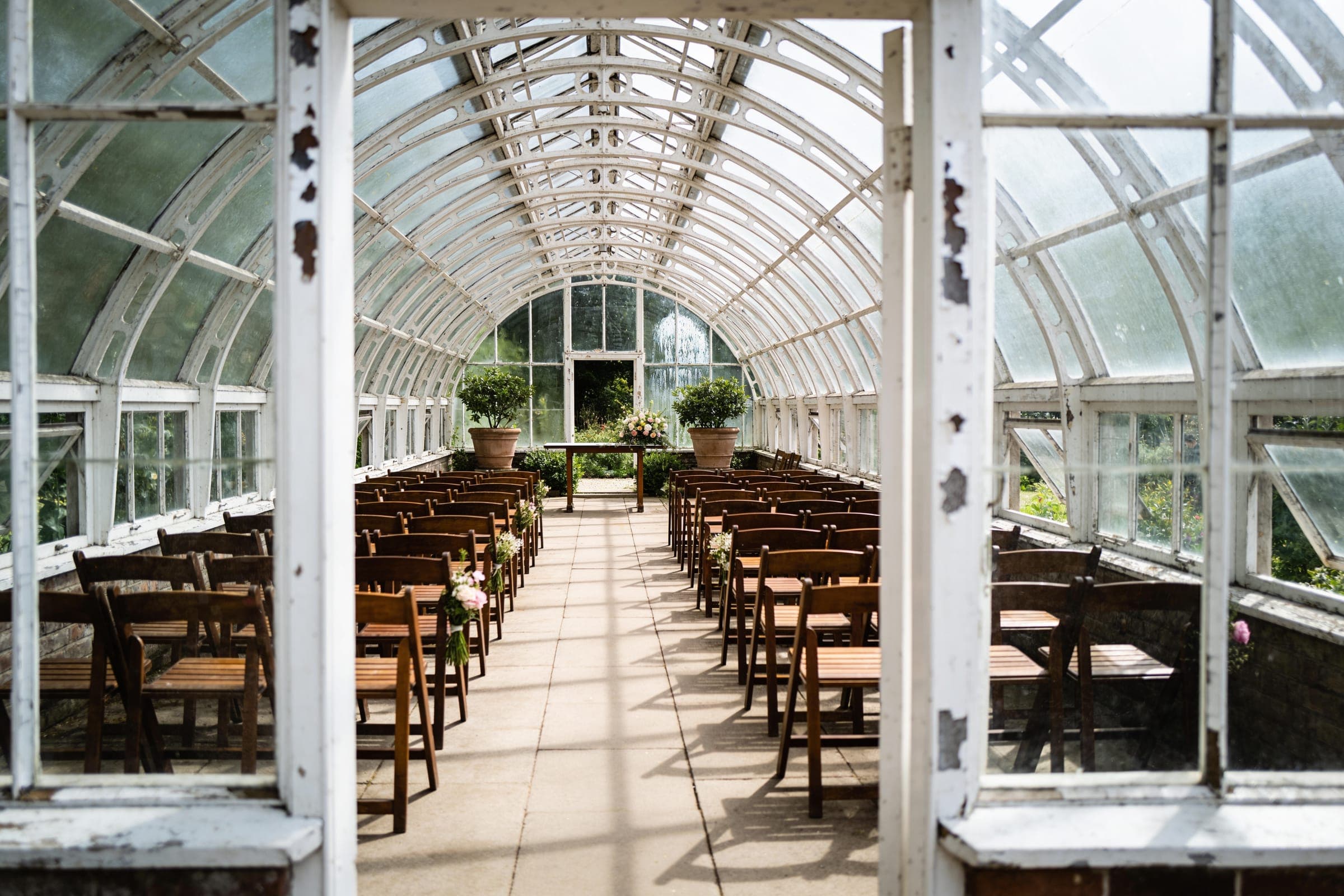 The central aisle of a long glasshouse in daylight, white iron ribs curving overhead and rows of wooden chairs leading to a small ceremony table.