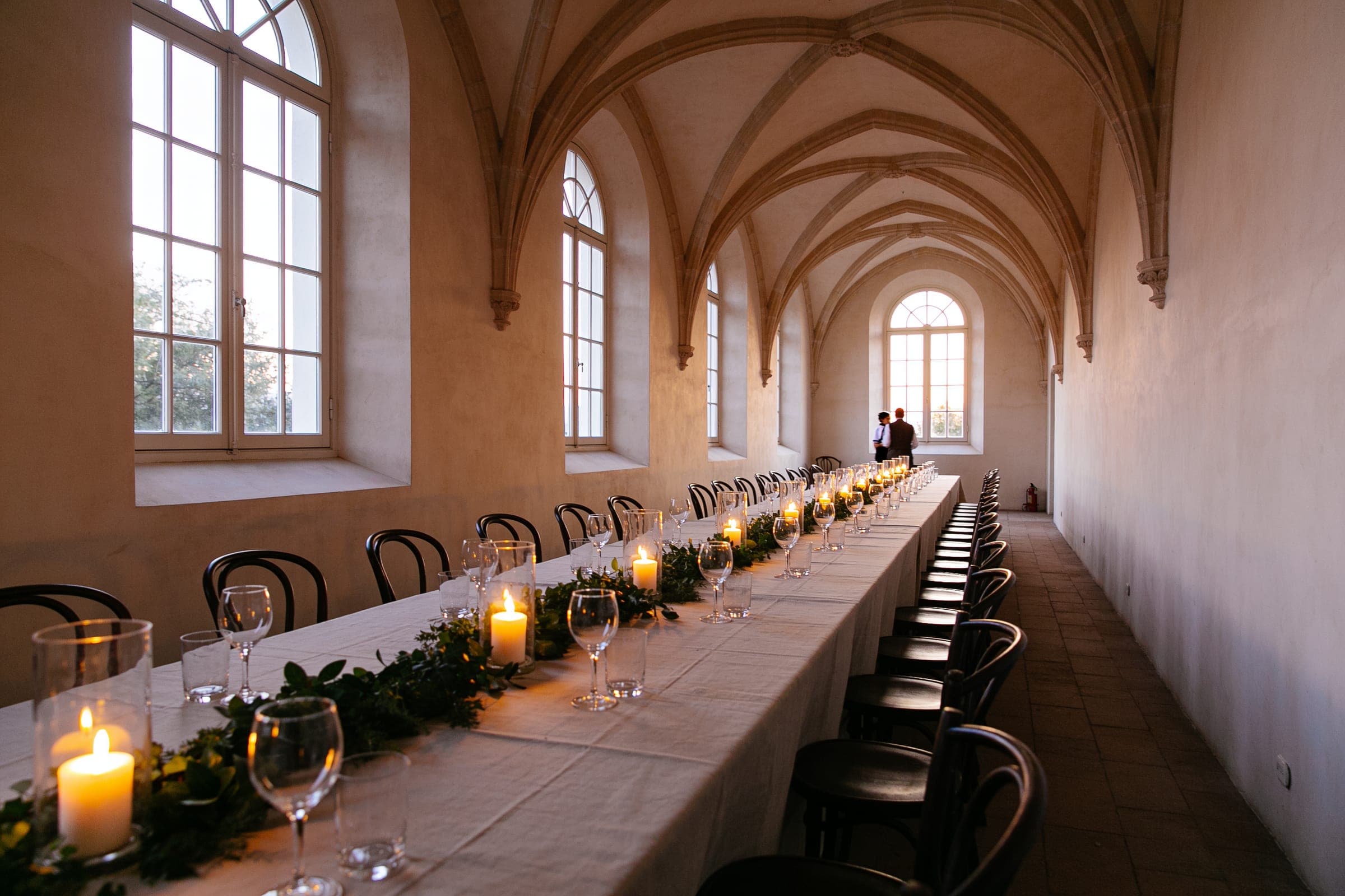 One long table with candles and greenery running the length of a narrow vaulted gallery lined with tall windows.