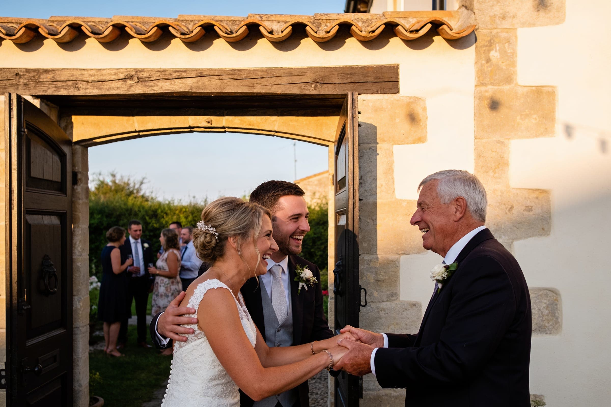 A couple laughing as they greet an older guest at a garden gateway, other guests talking in the garden behind.