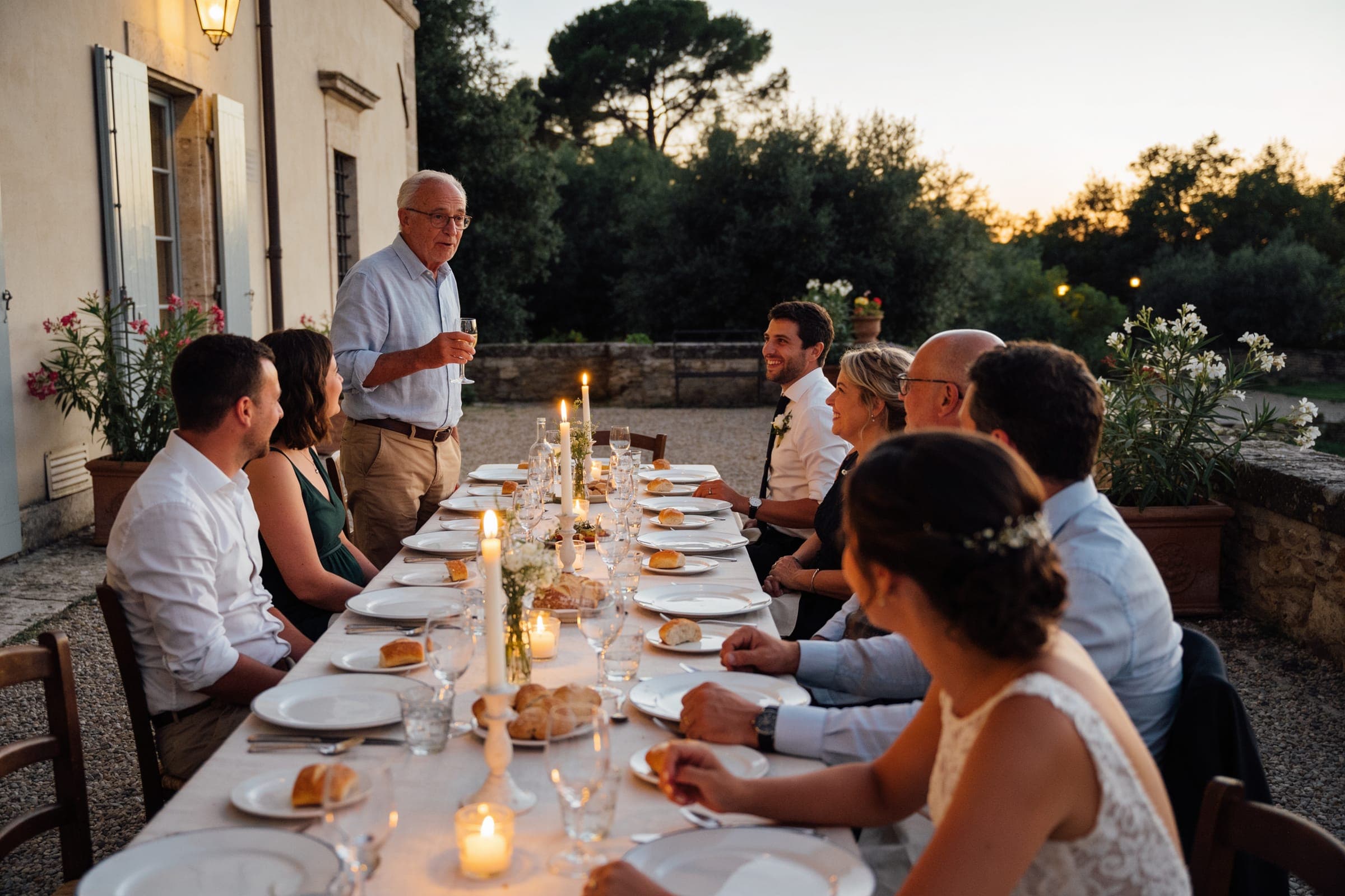 An older guest standing to speak at a small dinner on a terrace at dusk while the others listen around the table.