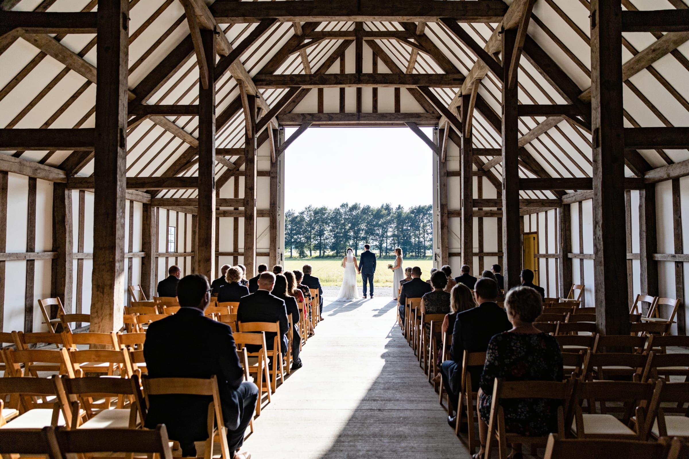 Guests seated along a timber barn facing a couple at its wide open end door, with fields and trees bright beyond.