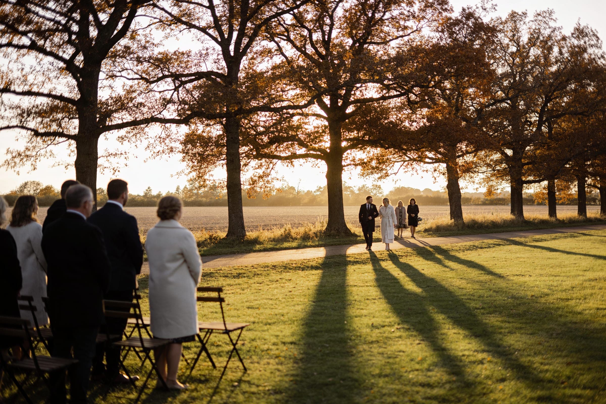A couple walking along a path under autumn trees in low sun, long shadows across the grass and guests standing by their chairs.