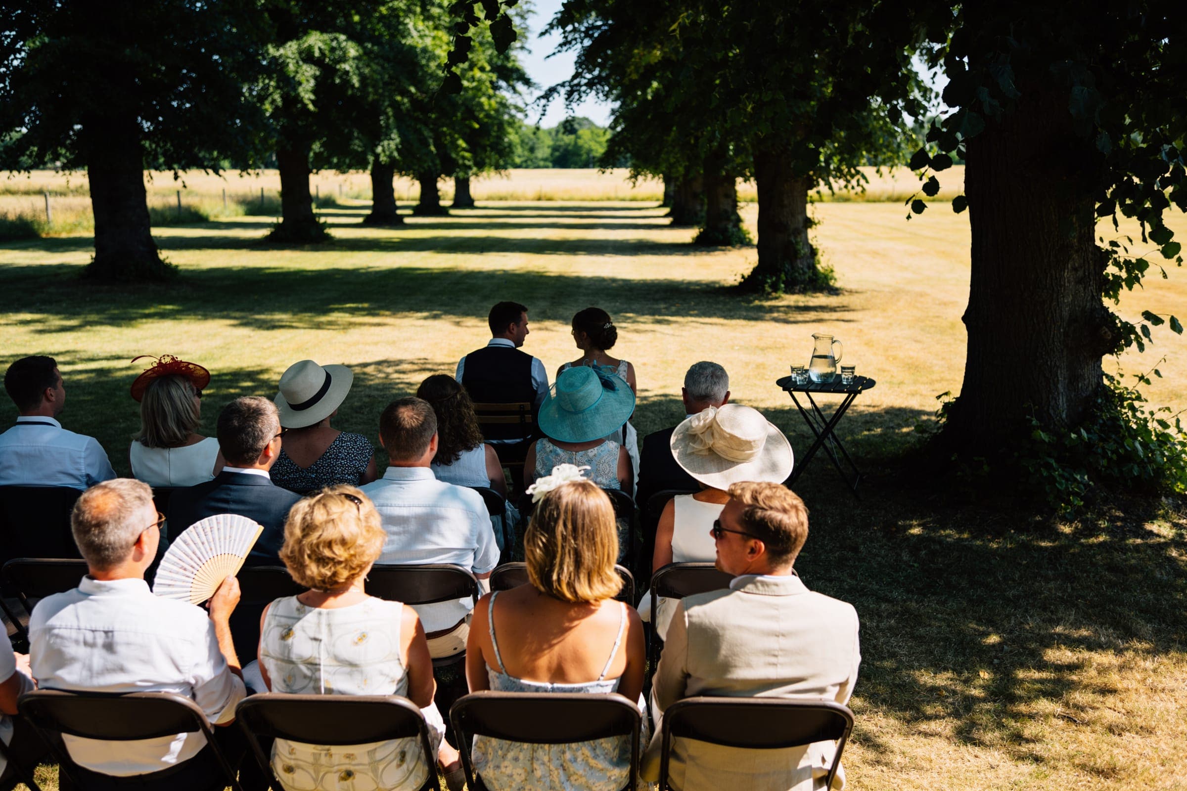 Guests in hats, one with a fan, seated in the deep shade of large trees at a summer ceremony, a table with a water jug nearby.