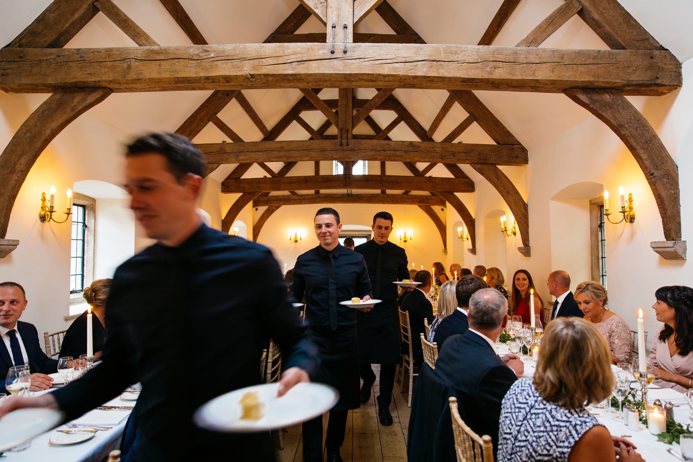 Waiting staff carrying plates between long tables of seated guests under a timber-beamed roof.