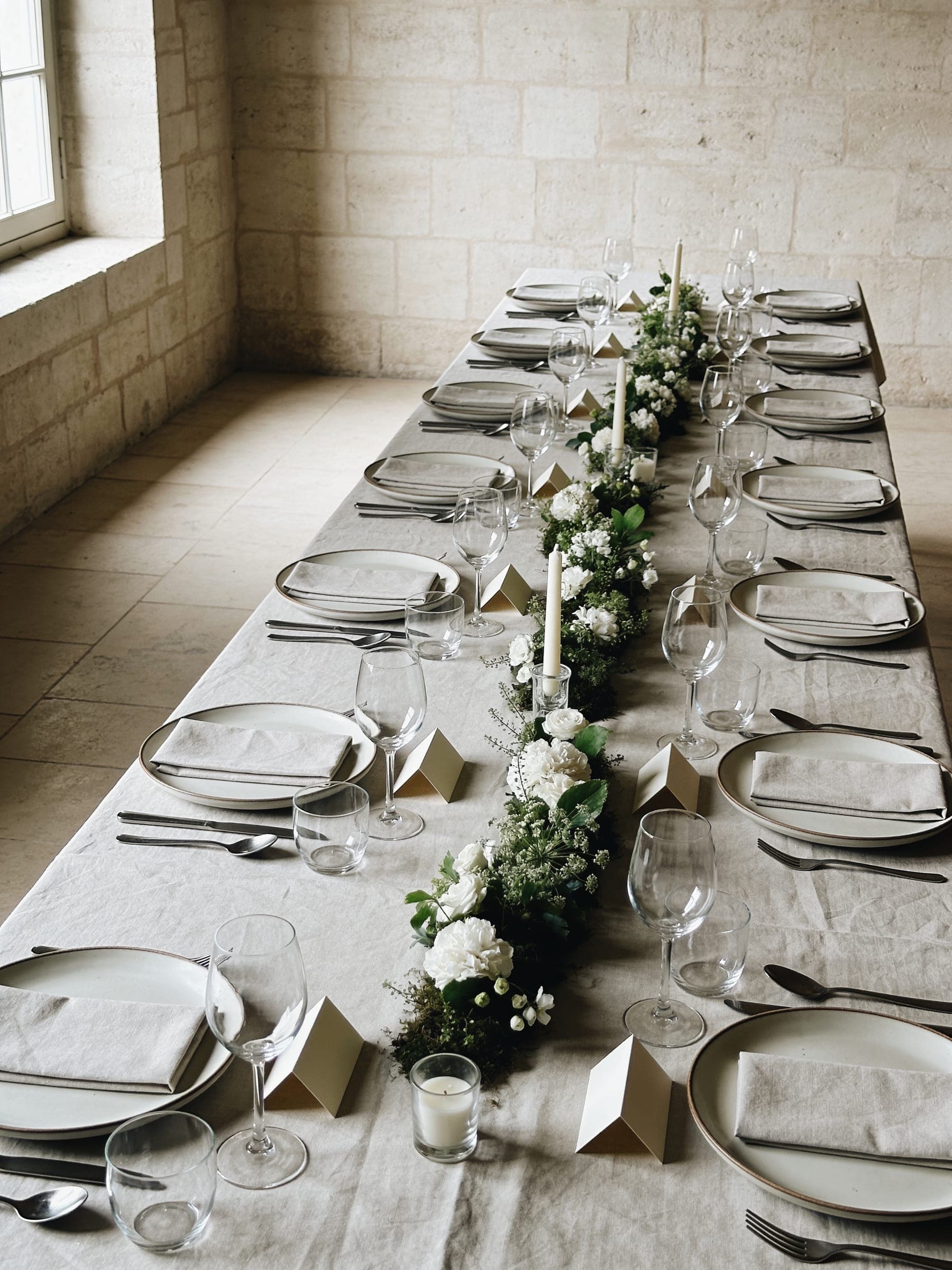 A long table in a pale stone room set with washed linen, matte plates, plain glasses, blank place cards and a low line of white flowers and candles.
