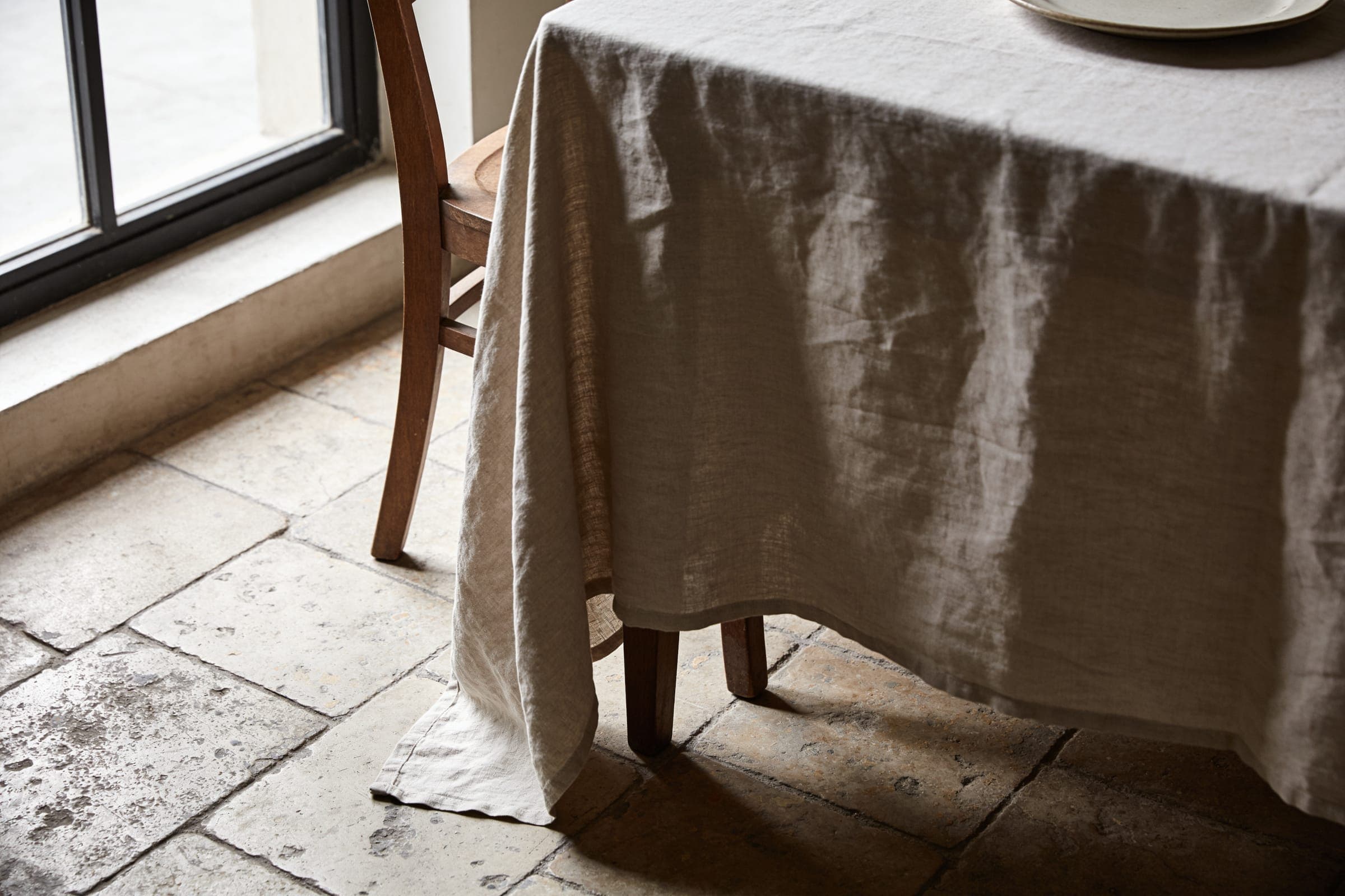 Washed oatmeal linen falling from a table corner to a worn stone floor, a wooden chair tucked beneath.