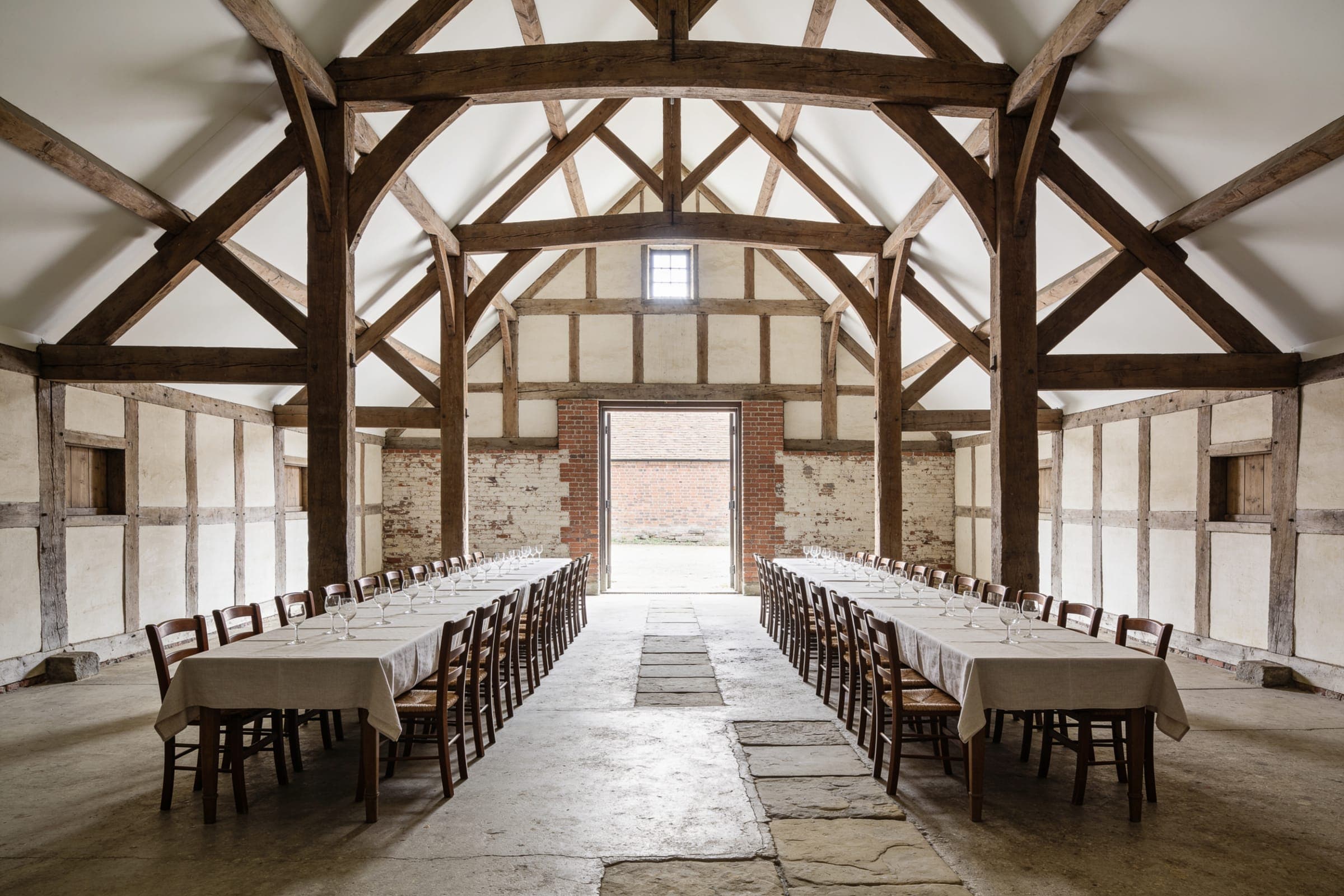 The interior of a tall timber-framed barn with exposed roof trusses and two long tables set along its length.