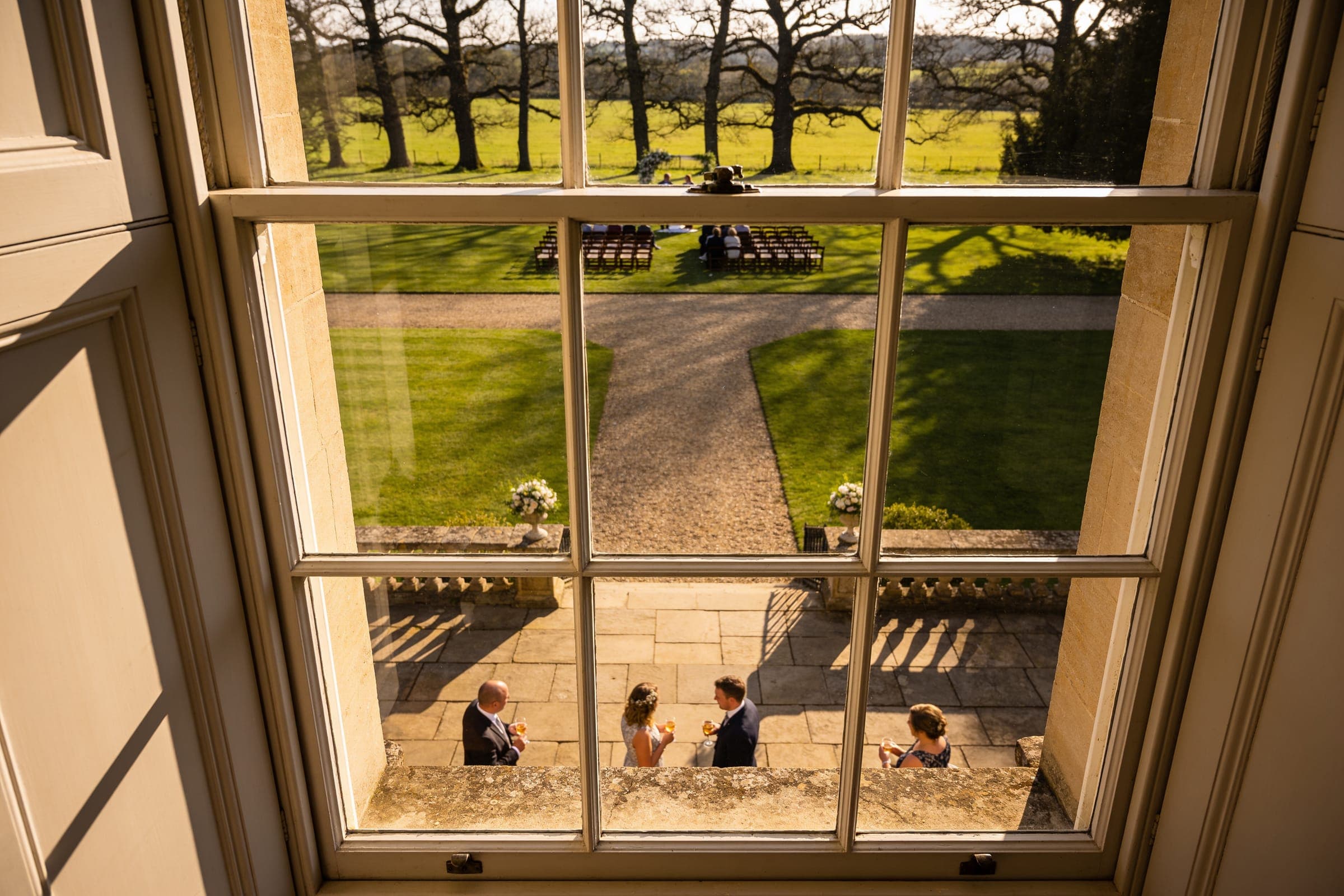 The view from an upper window of a country house: guests with drinks on the terrace below, a gravel path across the lawn and ceremony chairs set by distant trees.