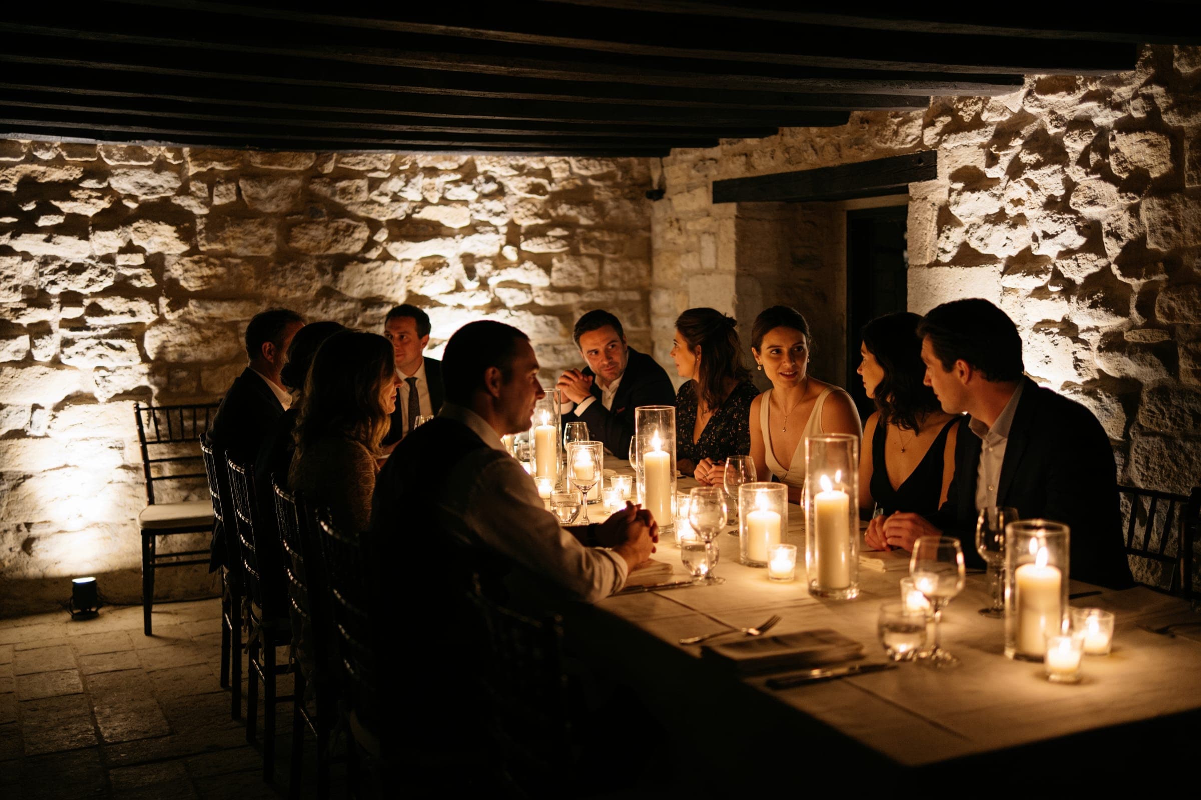 Guests at a table lit only by candles in a room with rough stone walls and a low timber ceiling.