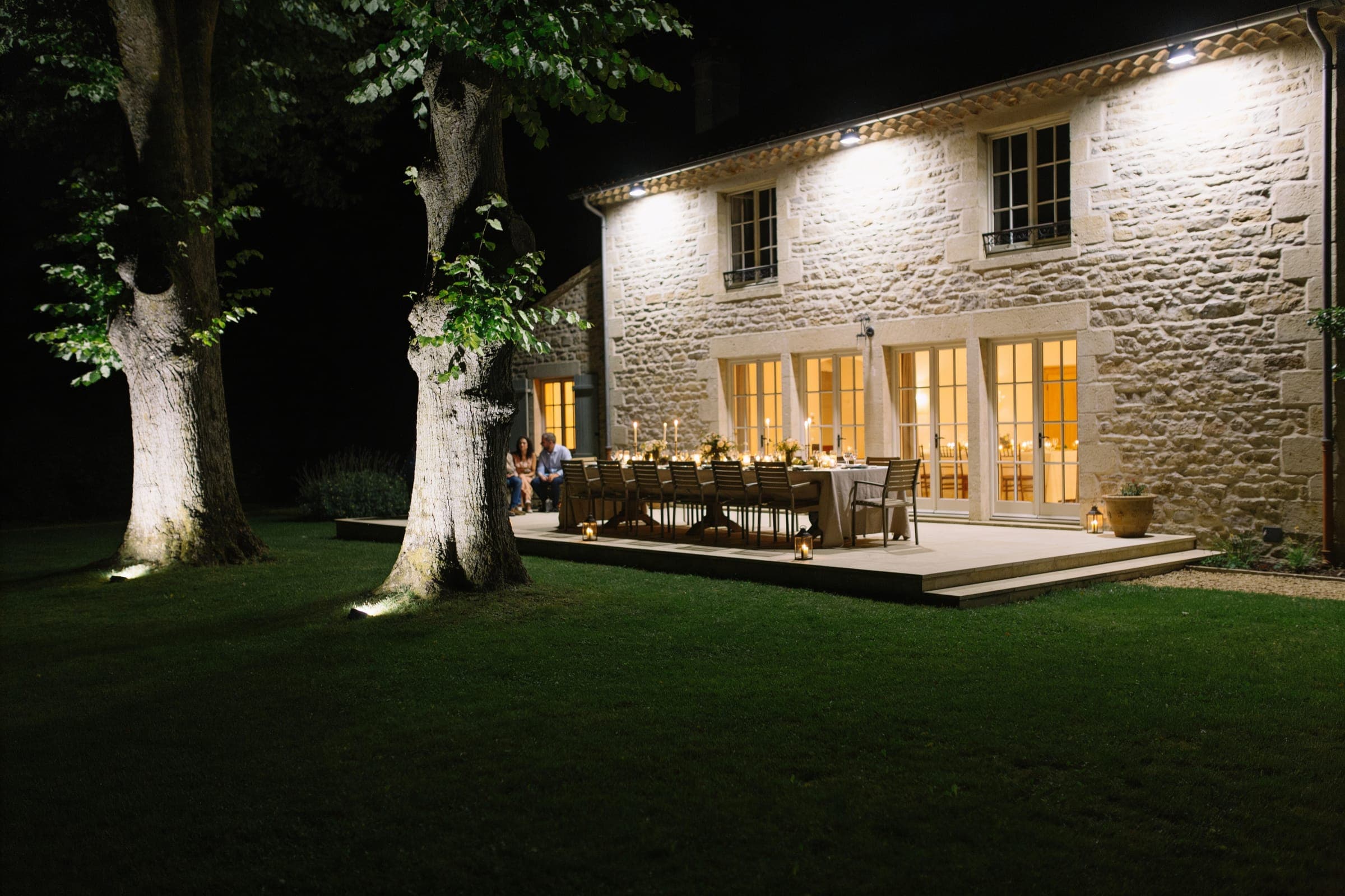 A dinner table on a terrace close to a stone house at night, its windows lit and two large trees lit softly from below.