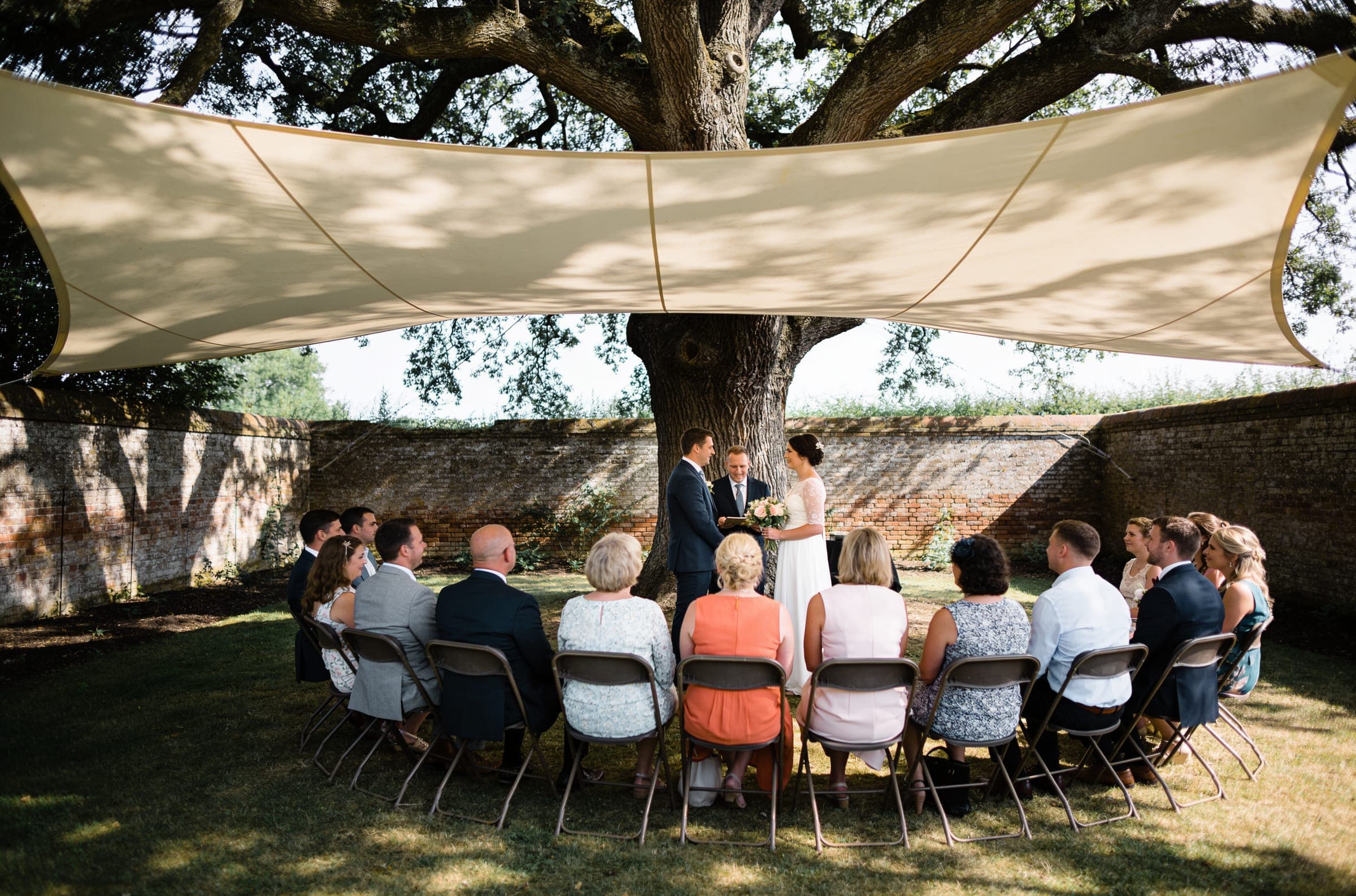 About twenty guests seated close in a semicircle under an old tree in a walled garden, around a couple and a celebrant.