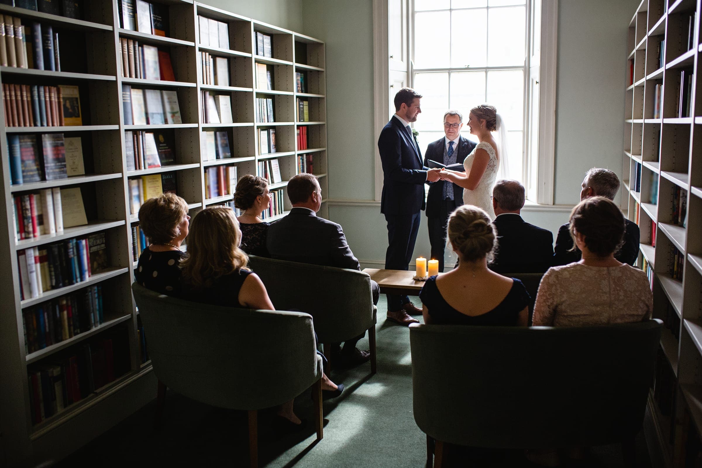 A couple exchanging vows in front of a tall window in a small book-lined room, a few guests seated close in armchairs.