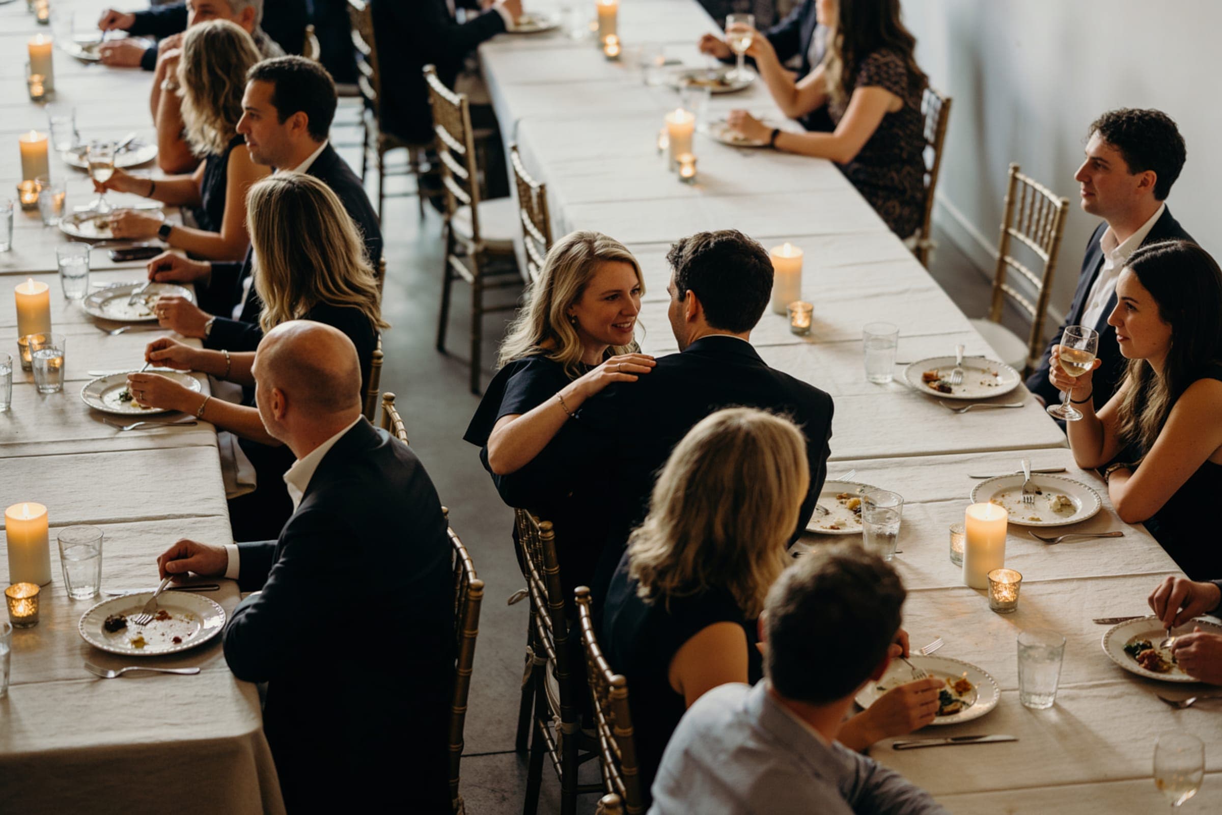 Guests at long tables talking in small groups with the people beside and opposite them, candles among the plates.