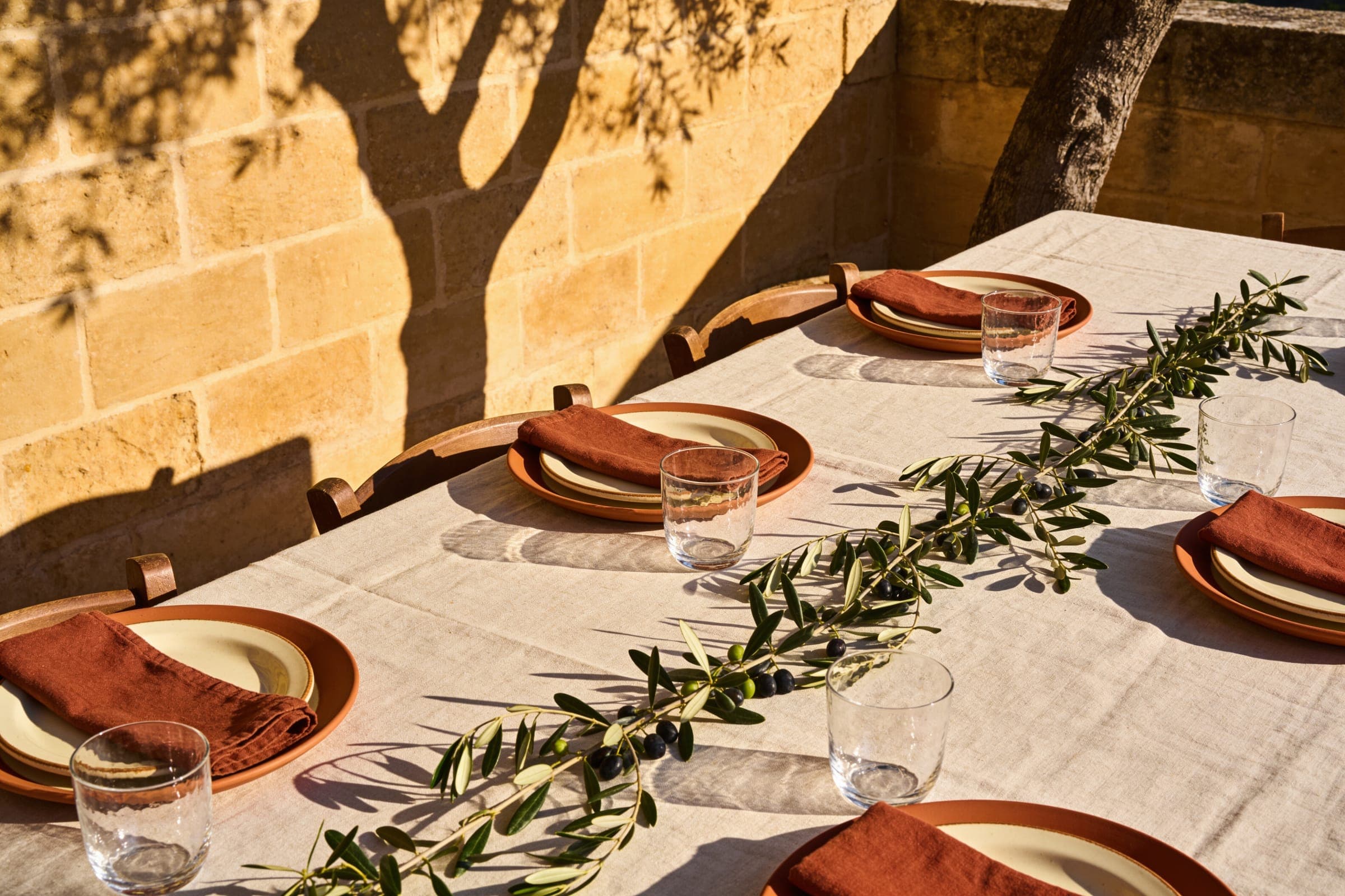 A table against a warm limestone wall with oatmeal linen, terracotta plates, rust napkins and an olive branch, a tree's shadow across the wall.