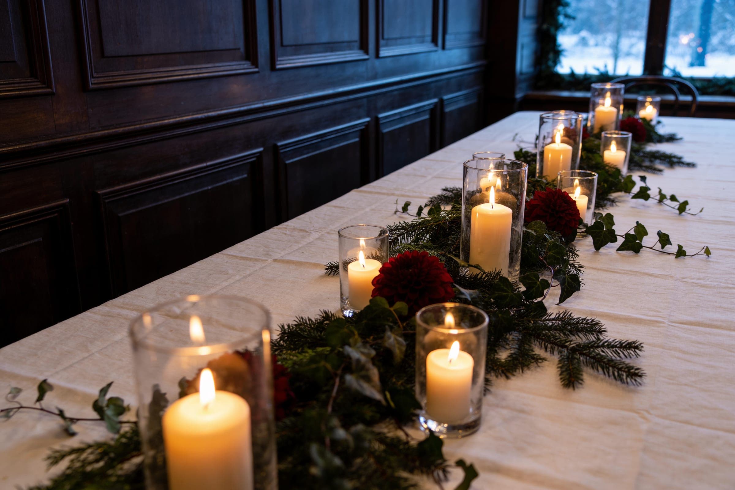 A white table with evergreen foliage, ivy, deep red flowers and candles in glass in a dark wood-panelled room, snow outside the window.