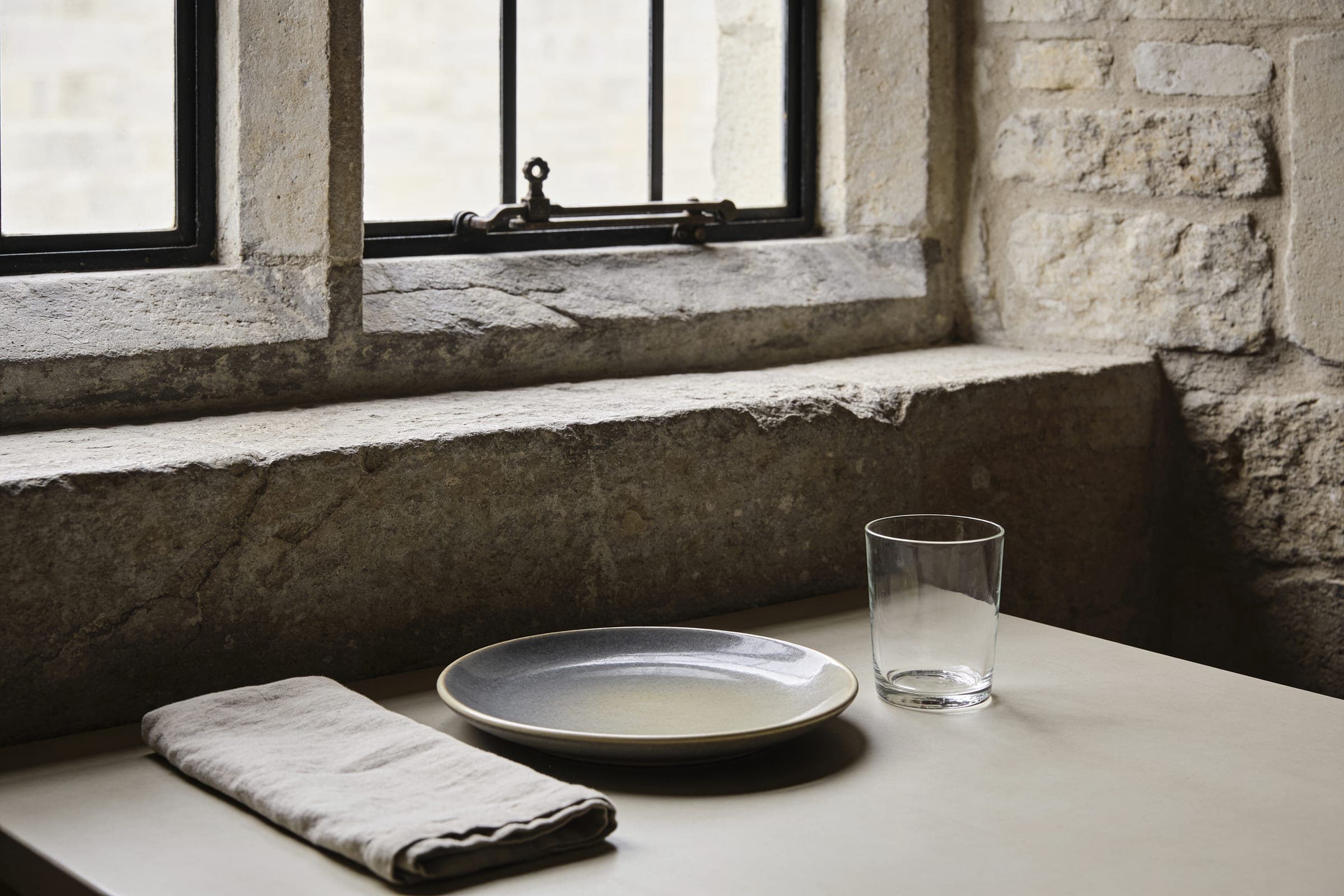 A gray-glazed plate, a plain glass and a linen napkin on a table beneath a deep, worn stone windowsill.