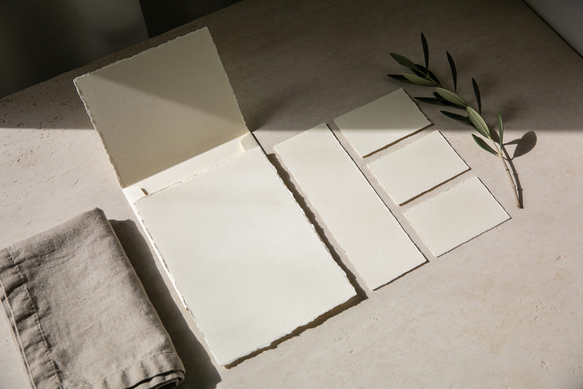 Blank cards in the same deckle-edged ivory paper laid on pale stone beside a linen napkin and an olive sprig.