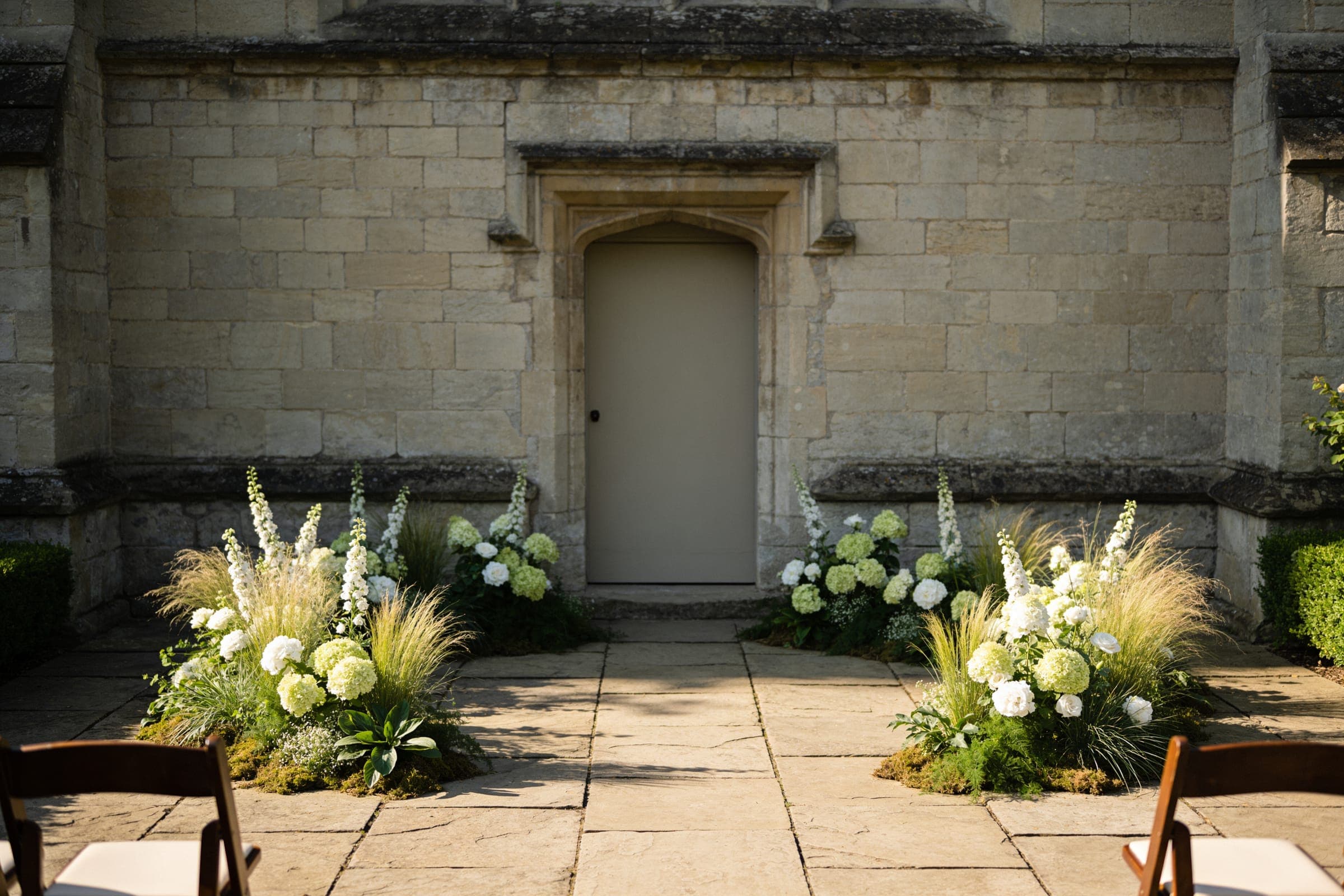 Low drifts of white flowers and grasses on the flagstones either side of a doorway in an old stone wall, with ceremony chairs at the edges.