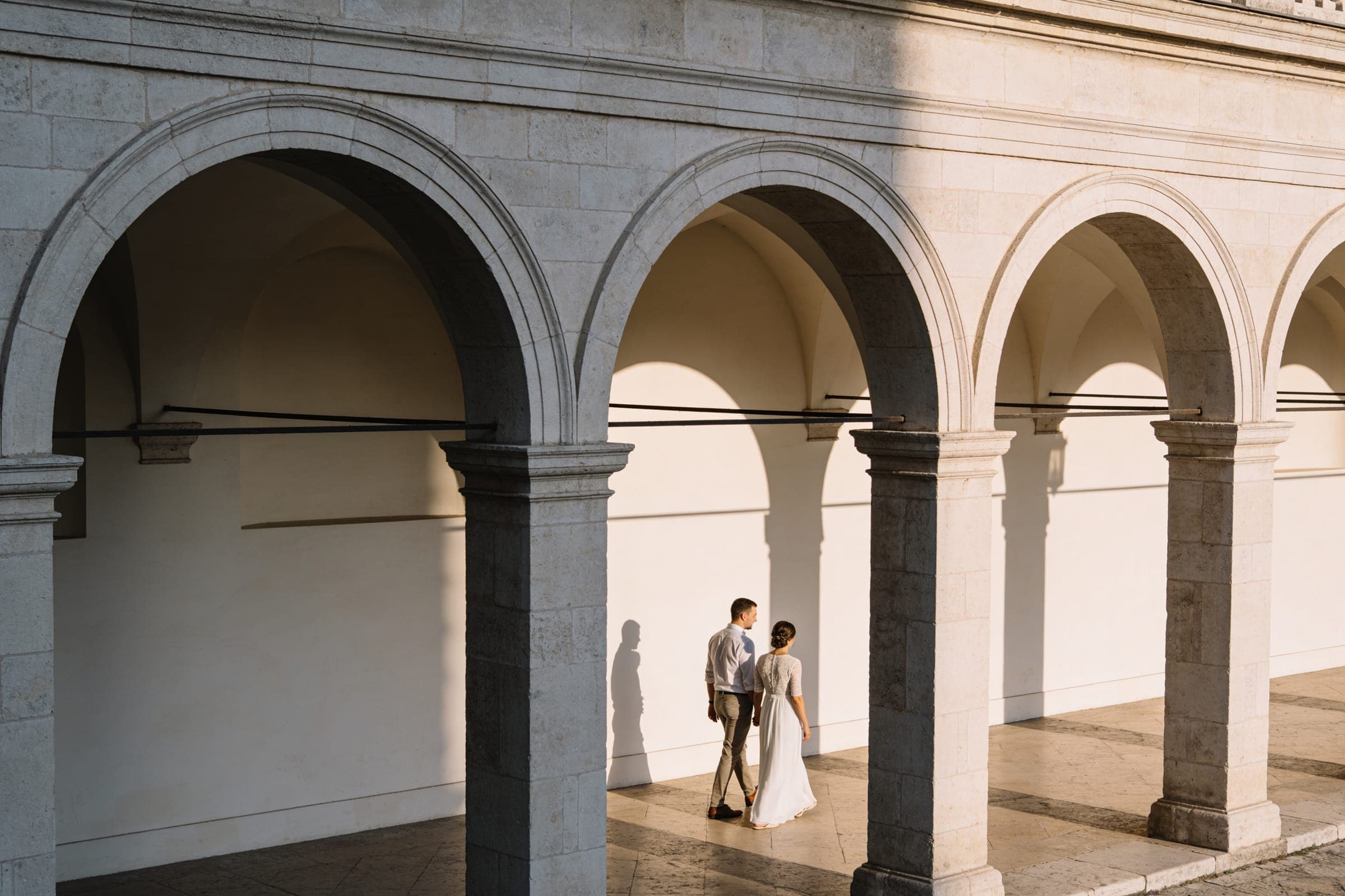 A couple walking through a pale stone arcade in late-afternoon light, small among the arches and their shadows.